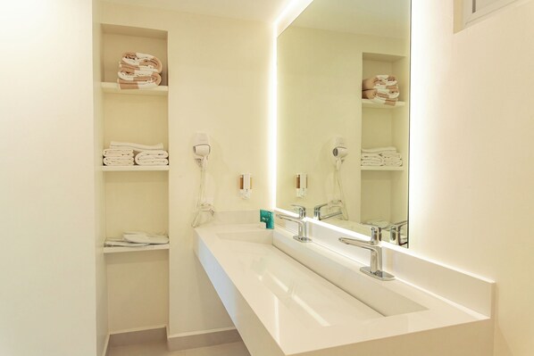 A modern bathroom with a white sink, two hair dryers, and shelves with folded towels.