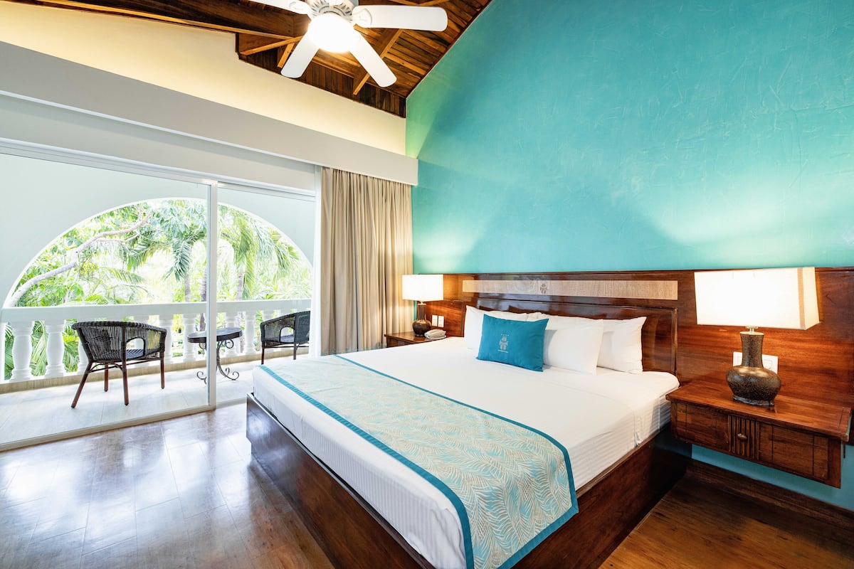 Une chambre d’hôtel avec un grand lit, une tête de lit en bois, une table de chevet avec une lampe, une vue sur l’extérieur grâce à une grande fenêtre et un balcon avec des chaises.