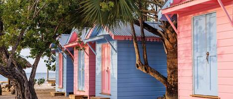 A row of beach huts with a sandy beach and palm trees in the background.