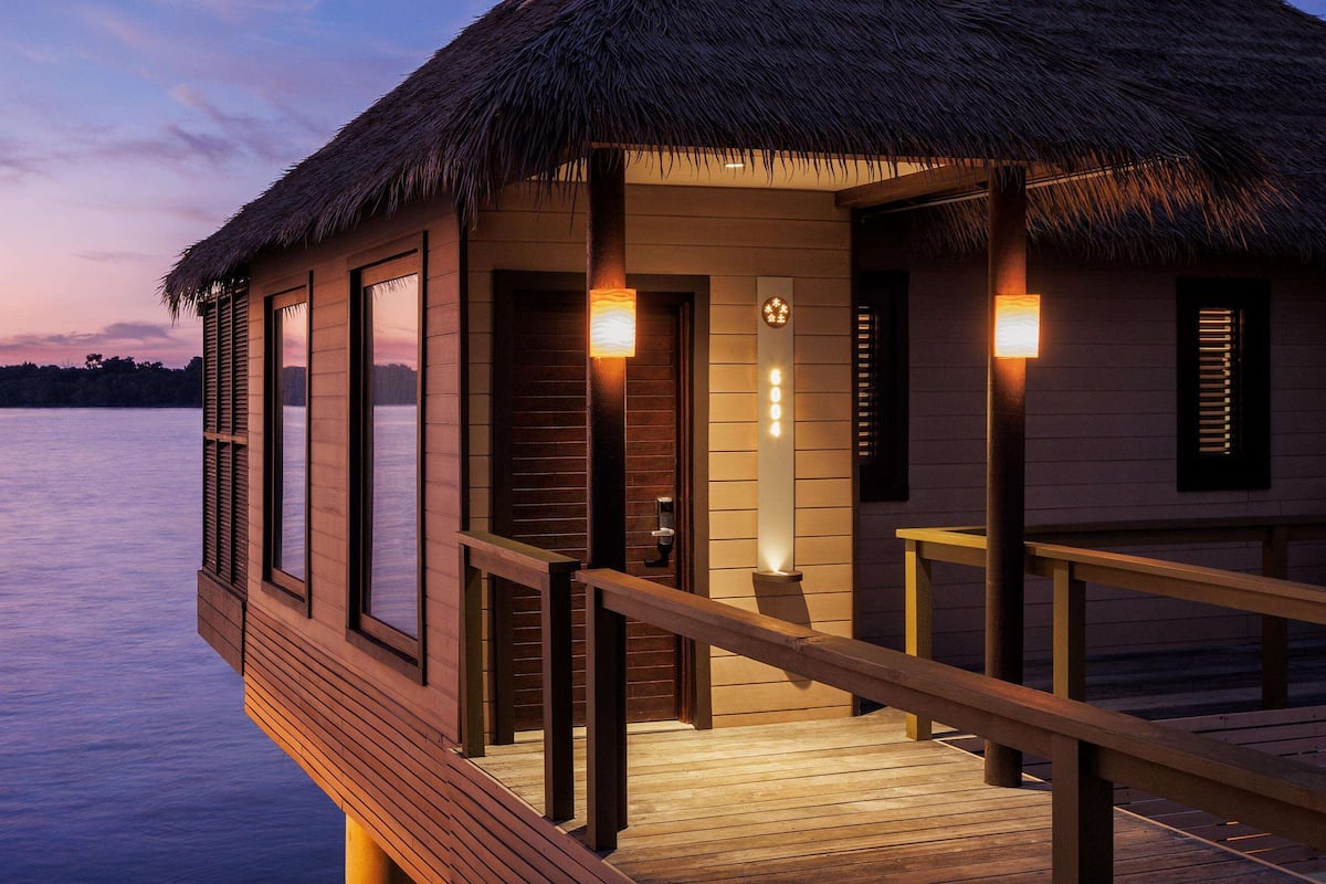 Een houten bungalow boven het water met rieten dak, houten leuningen en een houten terras.