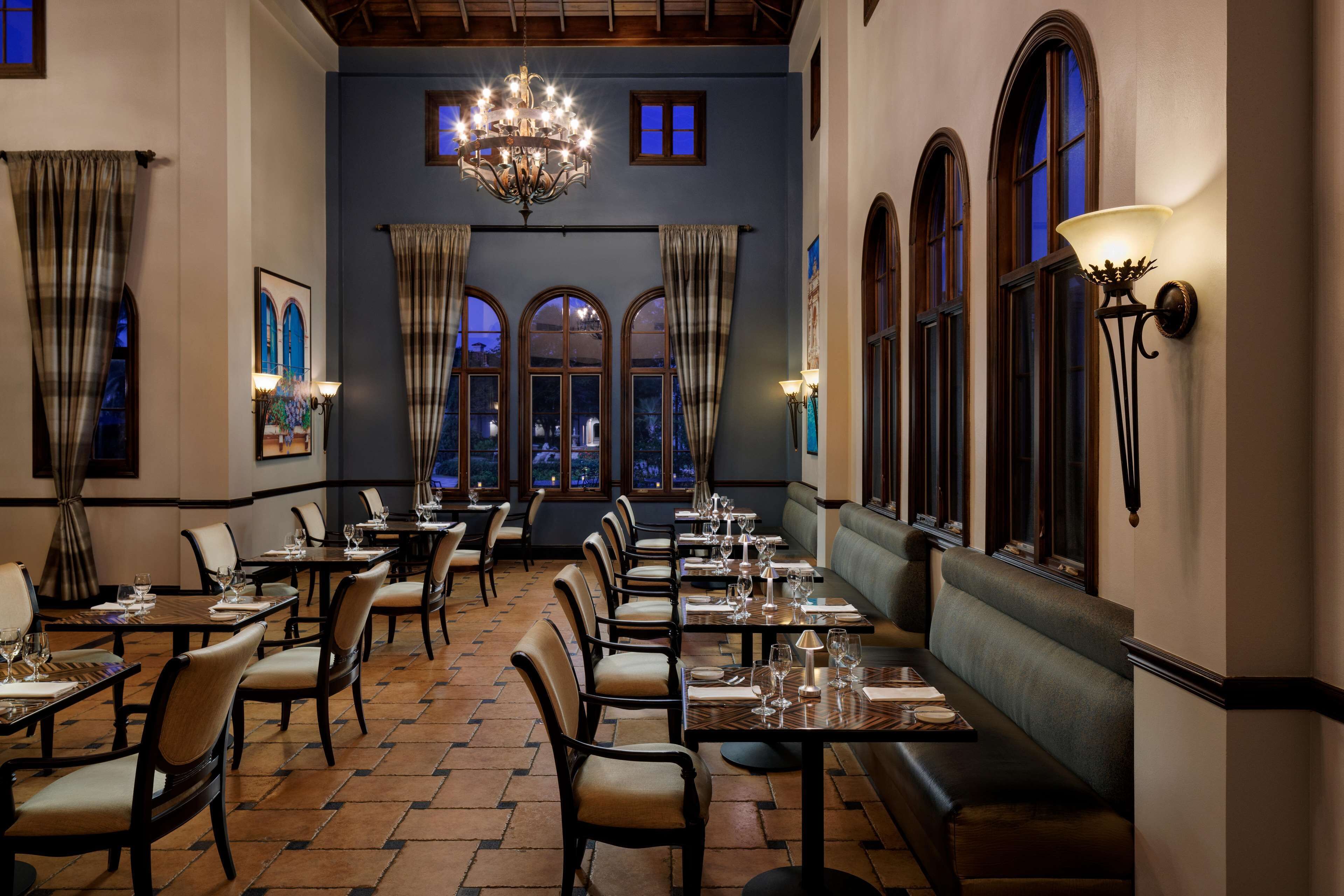 A restaurant with multiple tables and chairs, a chandelier, and large windows with curtains.