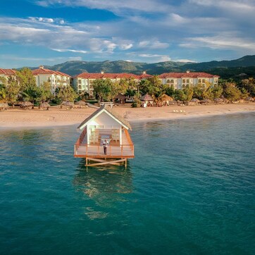 A wooden overwater bungalow with a balcony, surrounded by a beach and a row of buildings with red-tiled roofs.