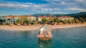 Un bungalow en bois construit sur pilotis, doté d’un balcon, entouré d’une plage et bordé d’une rangée de bâtiments aux toits recouverts de tuiles rouges.