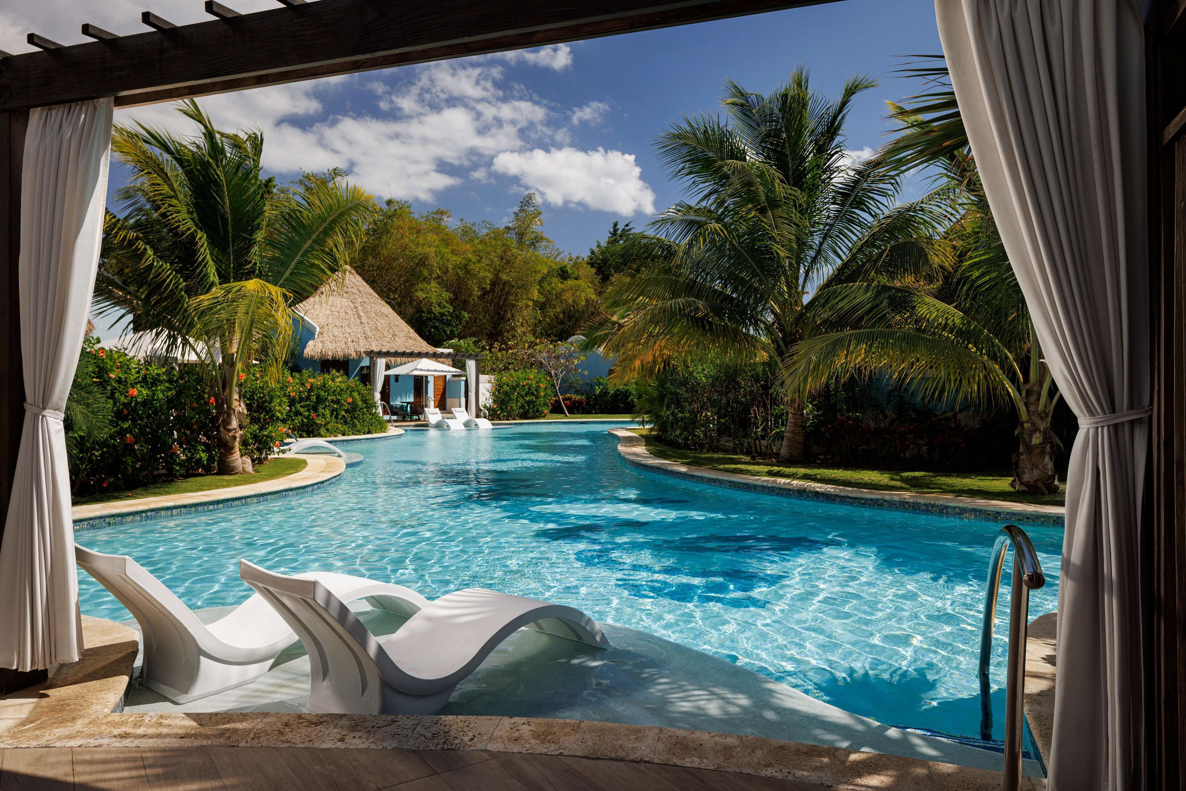 Une piscine bordée de chaises longues blanches, une structure à toit de chaume et des palmiers.
