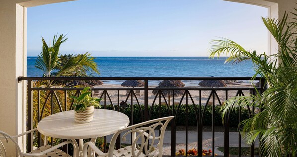 A balcony with a view of the beach, a round table with a potted plant, and white outdoor furniture.