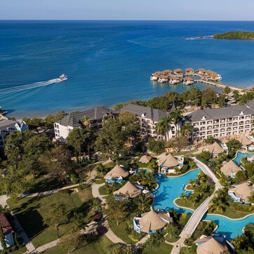 An aerial view of a resort with a large swimming pool, beachfront villas, and a boat dock.