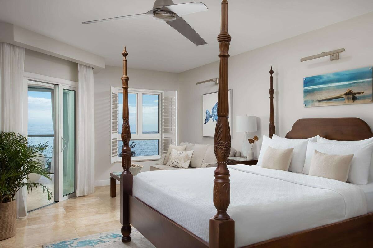 Een hemelbed met plafondventilator, een schilderij van een strand en uitzicht op de oceaan door een schuifdeur.