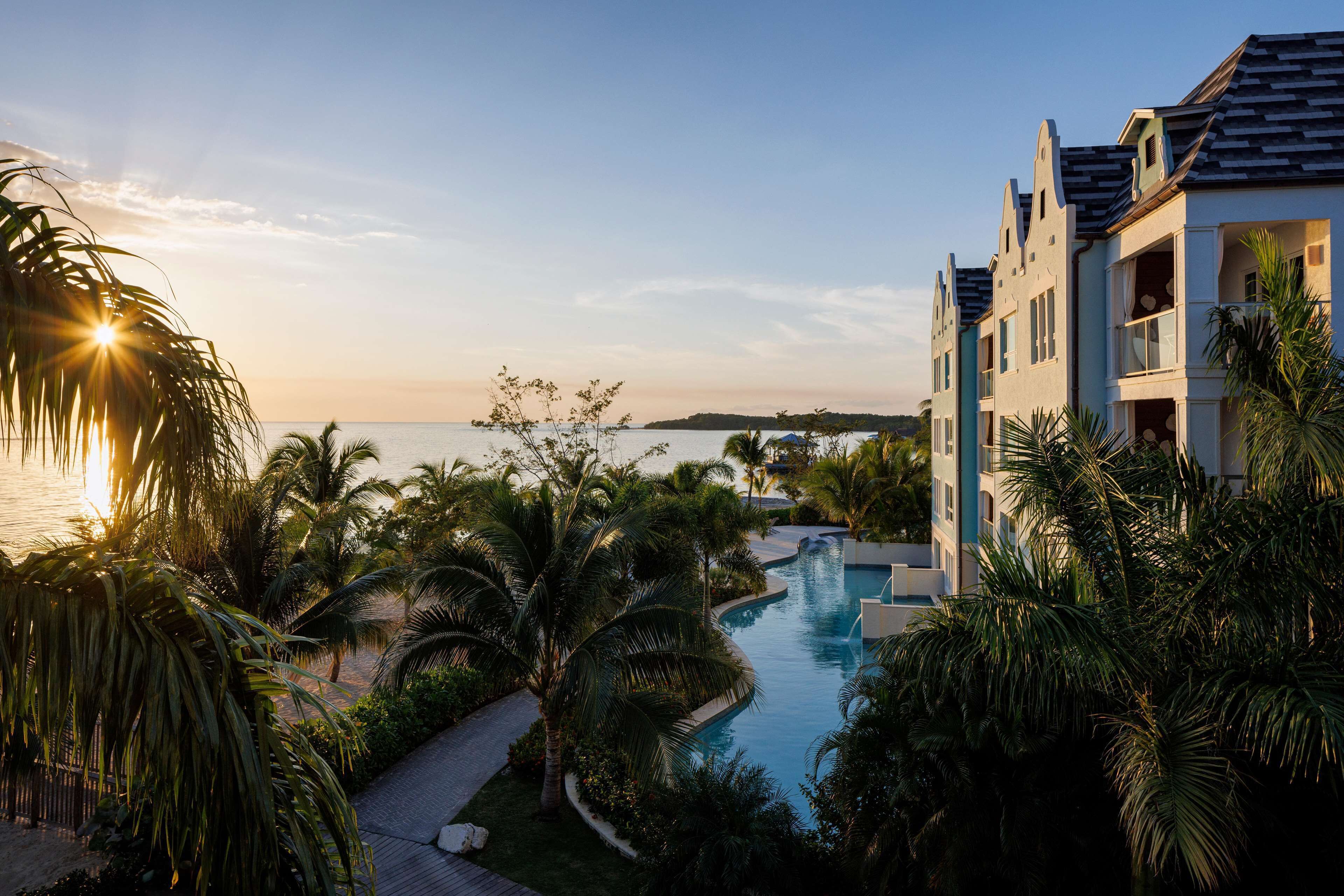 A resort with a swimming pool, palm trees, and a view of the sea at sunset.
