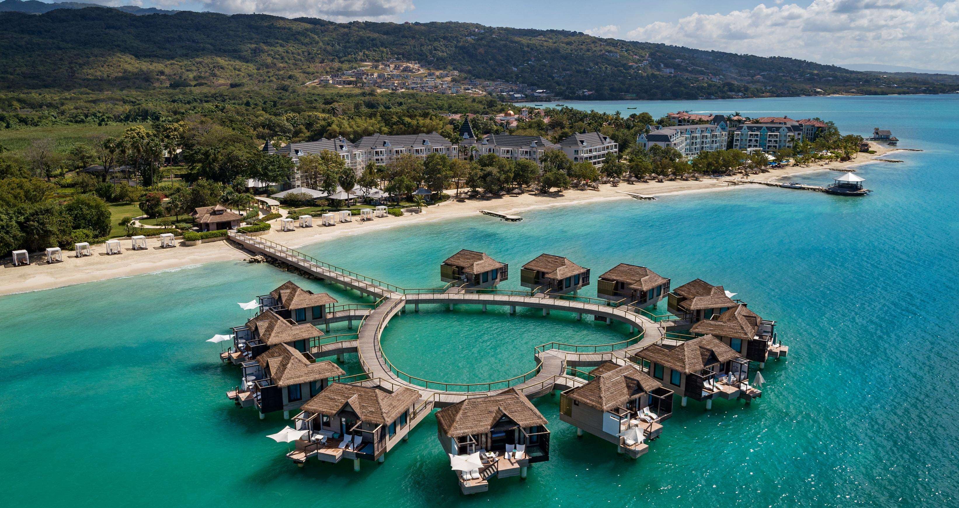 An overwater bungalows arrangement with a circular waterway connecting them, surrounded by clear turquoise waters and a beach.