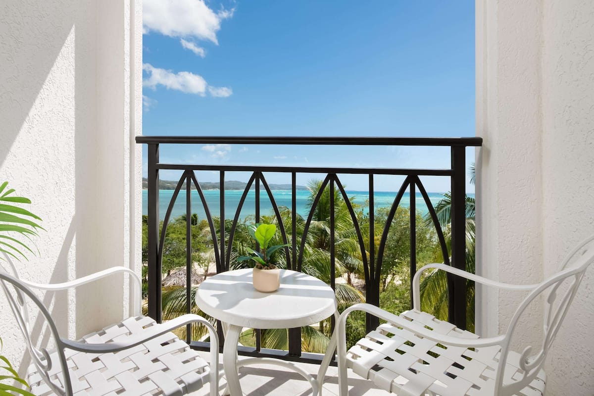Een balkon met witte stoelen en een kleine tafel, met uitzicht op een strand met palmbomen en de oceaan.
