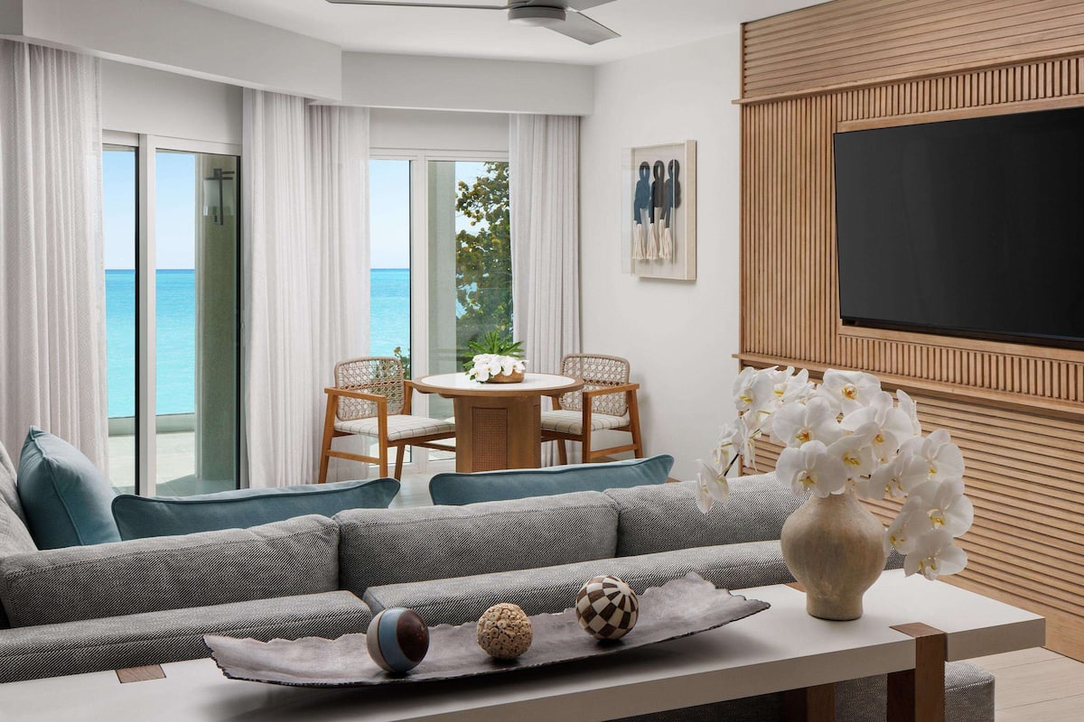 Een moderne woonkamer met een grijze bank, een eettafel met stoelen, een grote televisie en uitzicht op de oceaan.