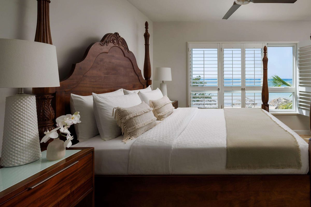 Een slaapkamer met een houten hemelbed, wit beddengoed, een nachtkastje met een lamp, een dressoir en uitzicht op de oceaan door het raam.