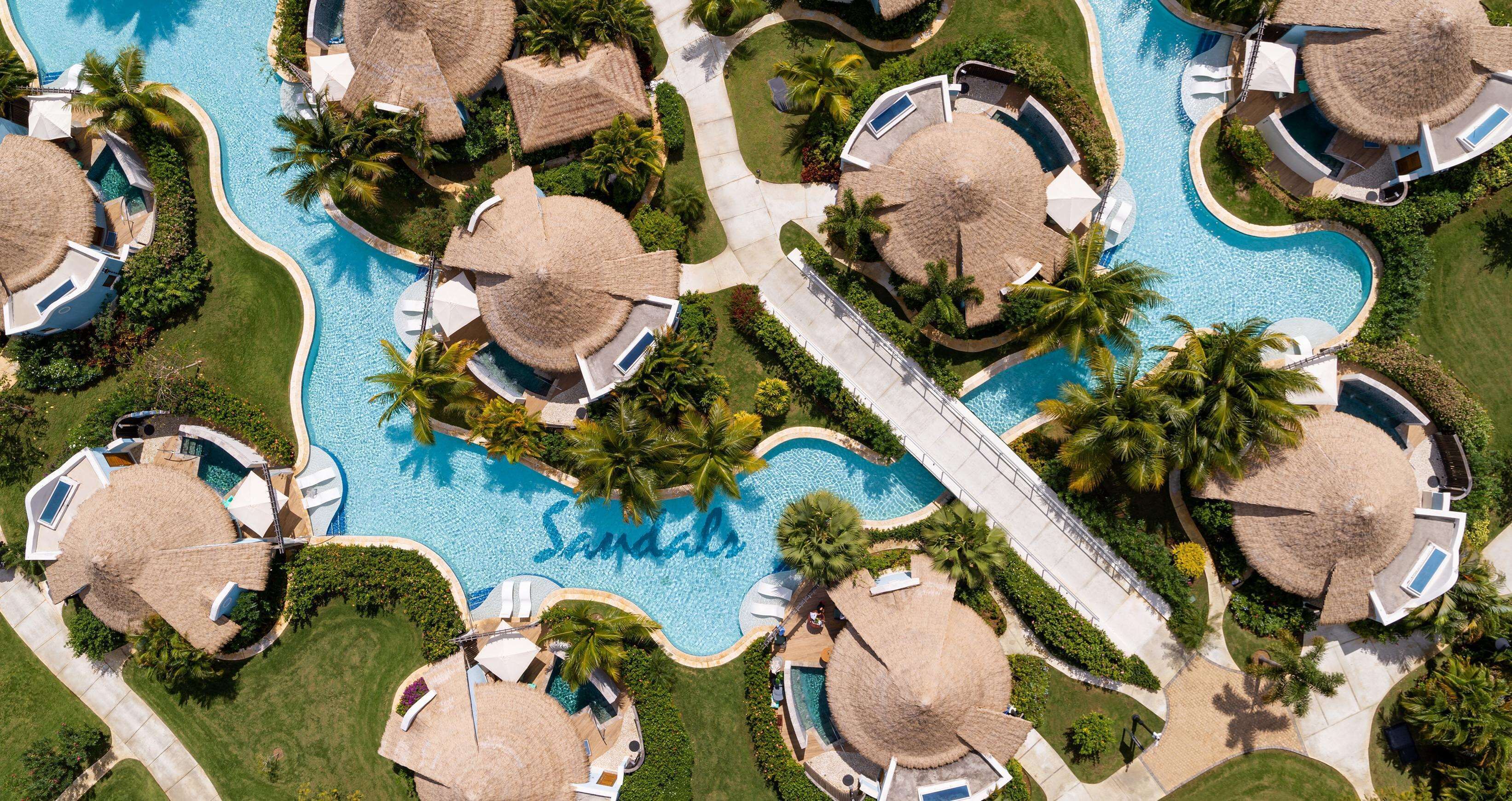 An aerial view of a resort with multiple thatched-roof villas, a central swimming pool, and lush greenery.