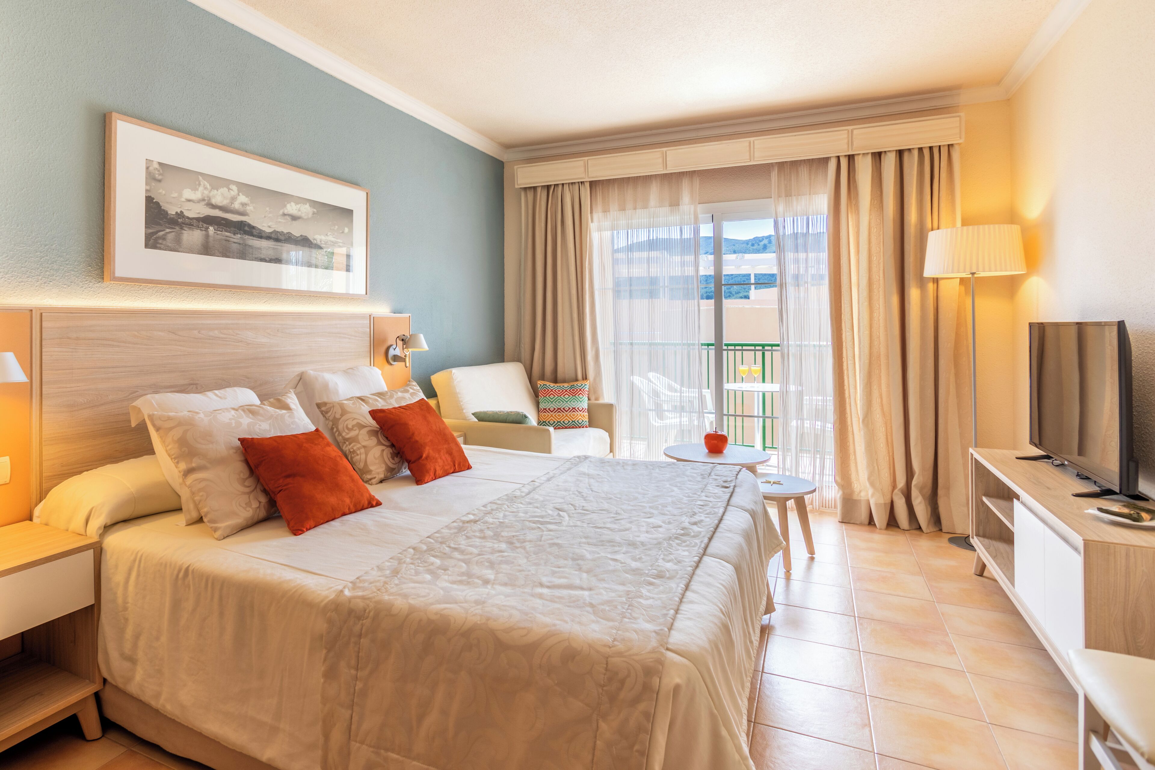 Habitación de hotel con una cama grande, televisión, una mesita y balcón con cortinas.