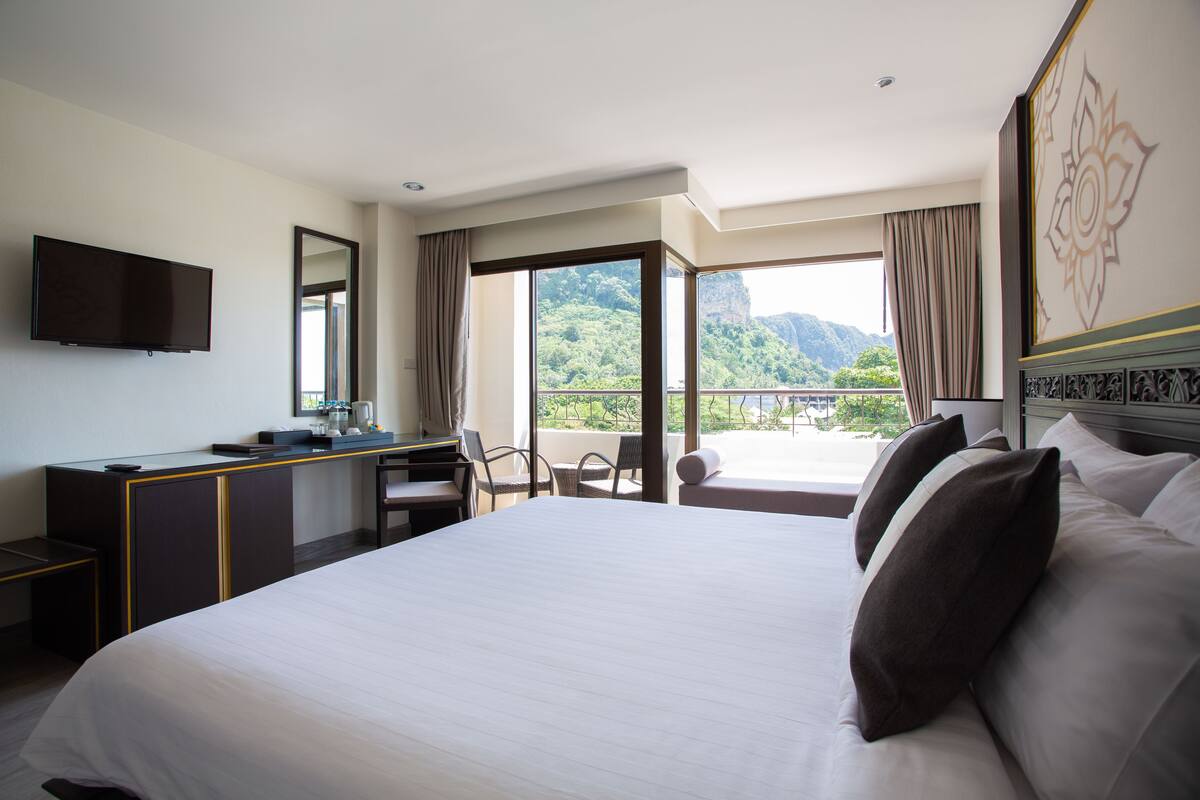 Ein Hotelzimmer mit einem großen Bett, einem Schreibtisch mit Flachbildfernseher, einem Sessel und einem Balkon mit Blick auf Grünflächen und Berge.