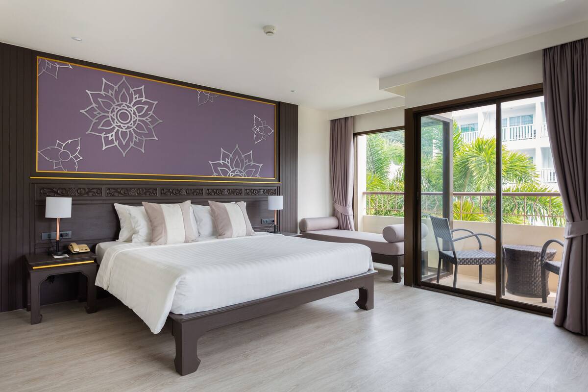 Ein modernes Hotelzimmer mit einem großen Bett, einem Nachttisch, einem Sessel und einem Balkon mit Blick auf die grüne Umgebung.