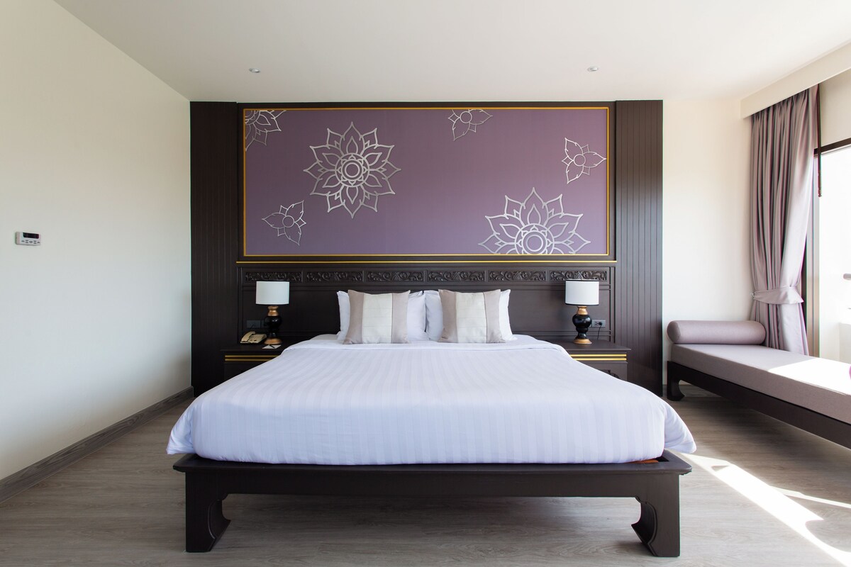 A hotel room with a large bed, a headboard with floral patterns, two bedside tables with lamps, and a window with curtains.