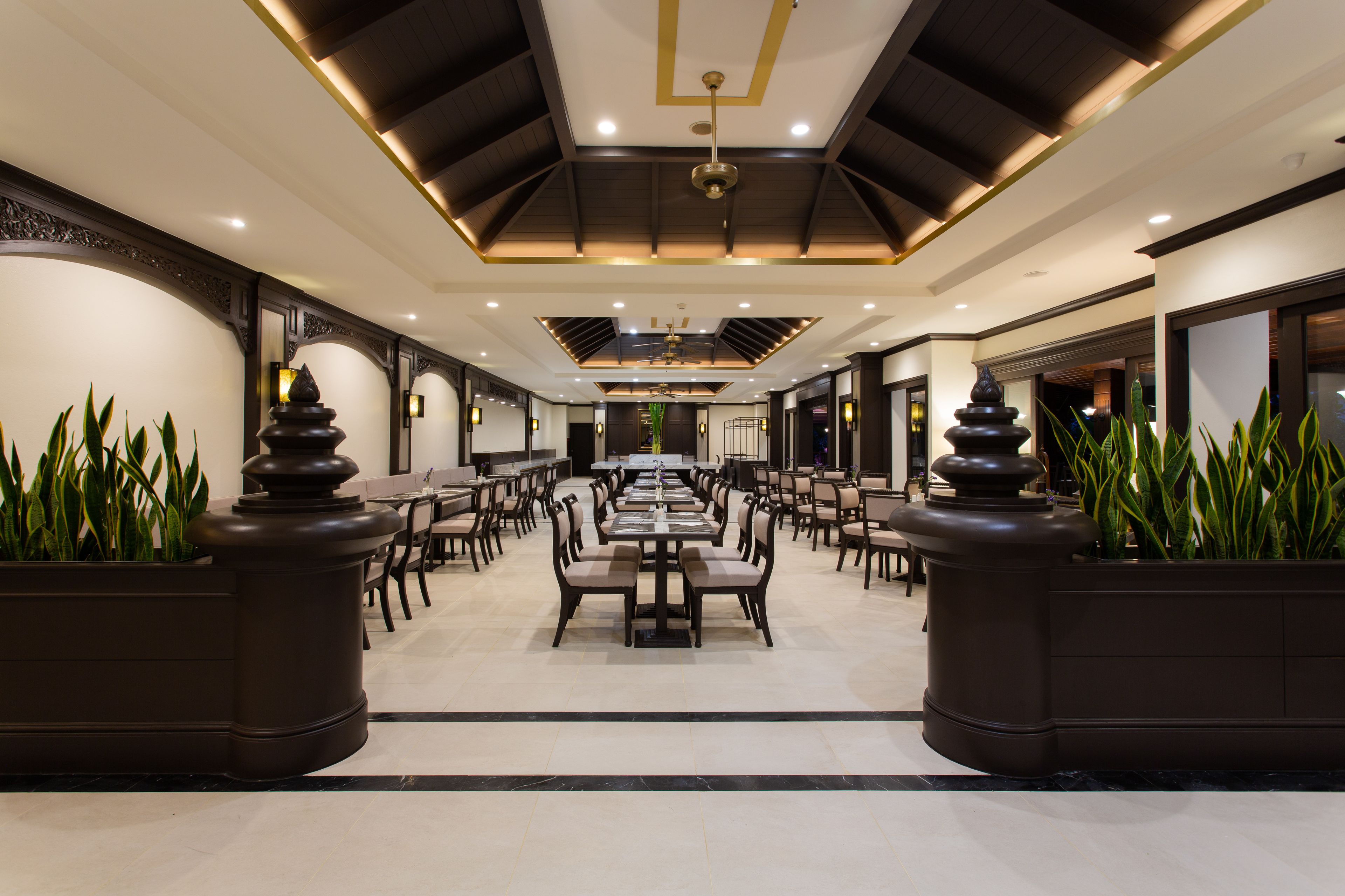 A spacious dining area with multiple tables and chairs, a high ceiling with recessed lighting, and decorative planters with green plants.