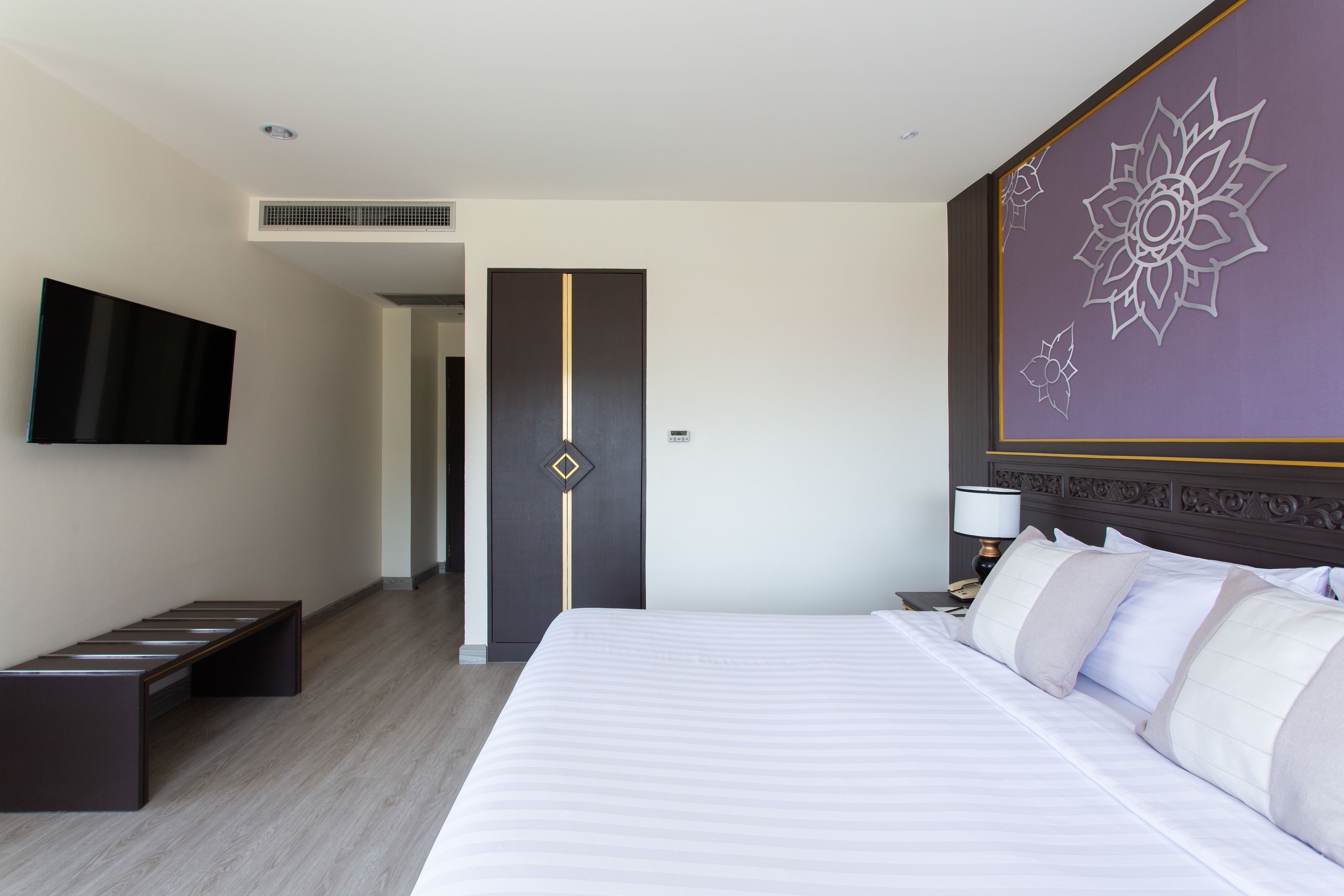 A hotel room with a large bed, a flat-screen TV mounted on the wall, a bench, and a decorative wall panel.