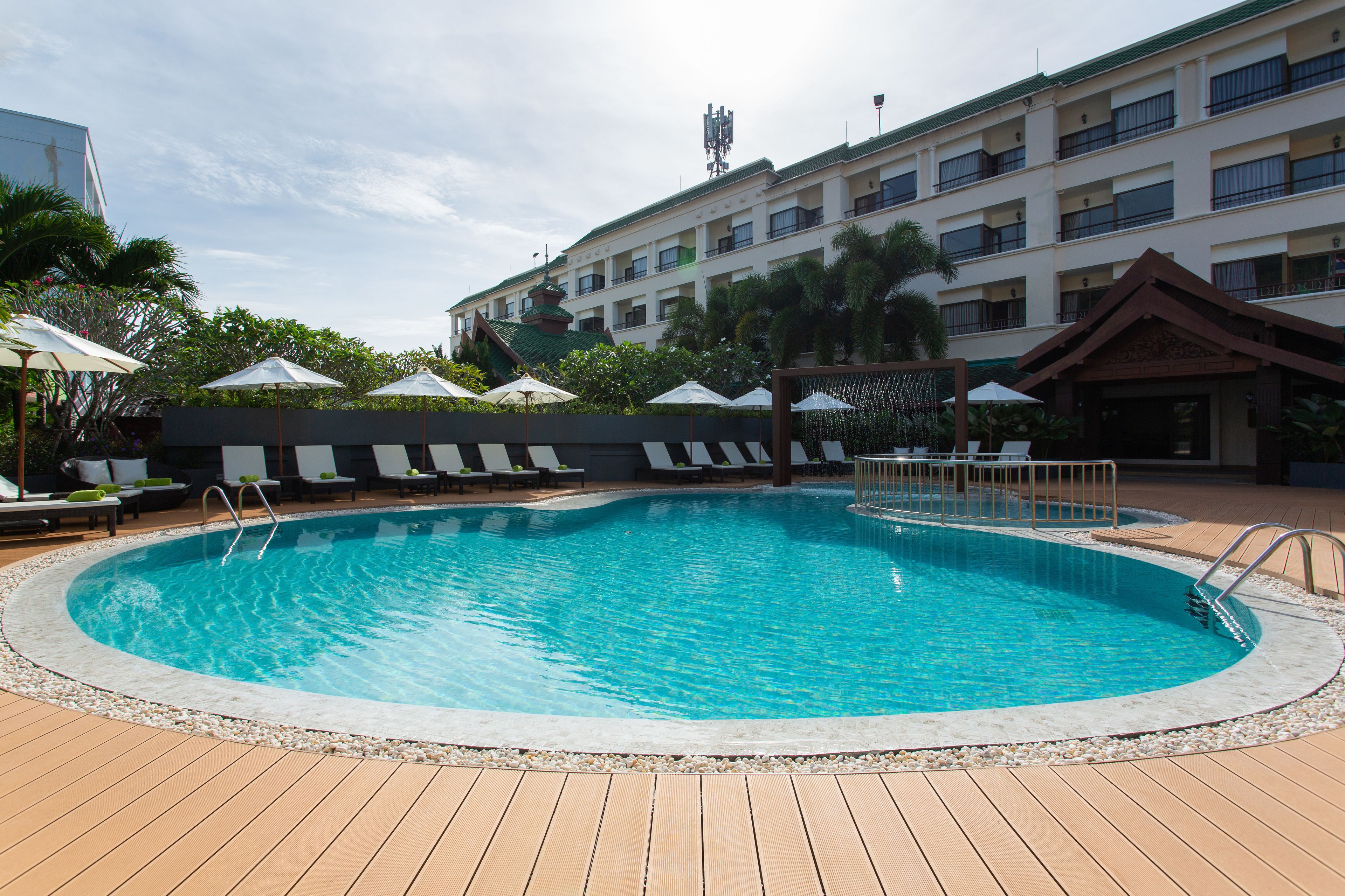 A swimming pool with lounge chairs and umbrellas, surrounded by a wooden deck and a building with balconies.