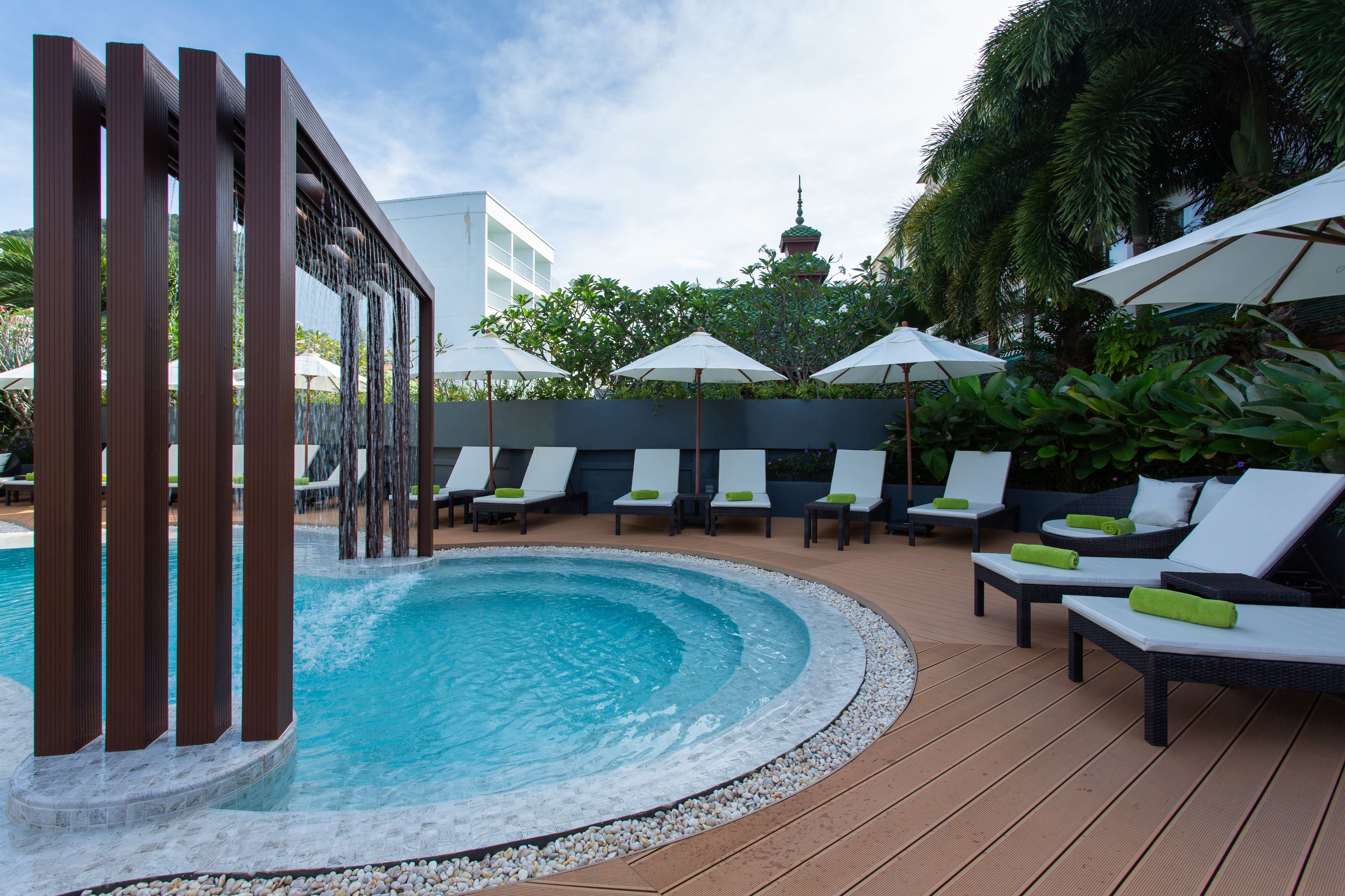 A pool area with lounge chairs and umbrellas, surrounded by greenery and a building with a tower.