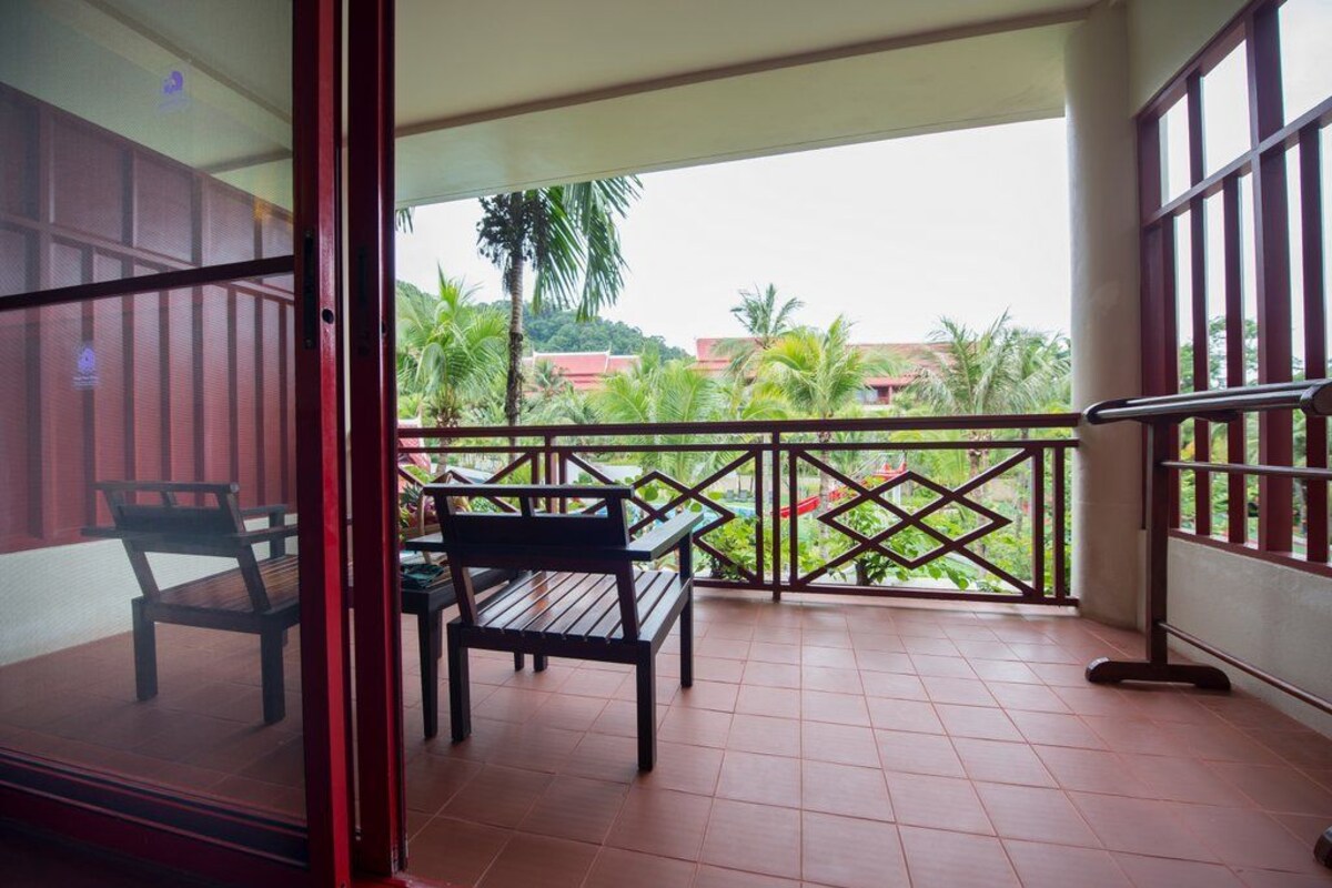 An open balcony with a wooden table and chairs, a metal railing, and a view of greenery and buildings.