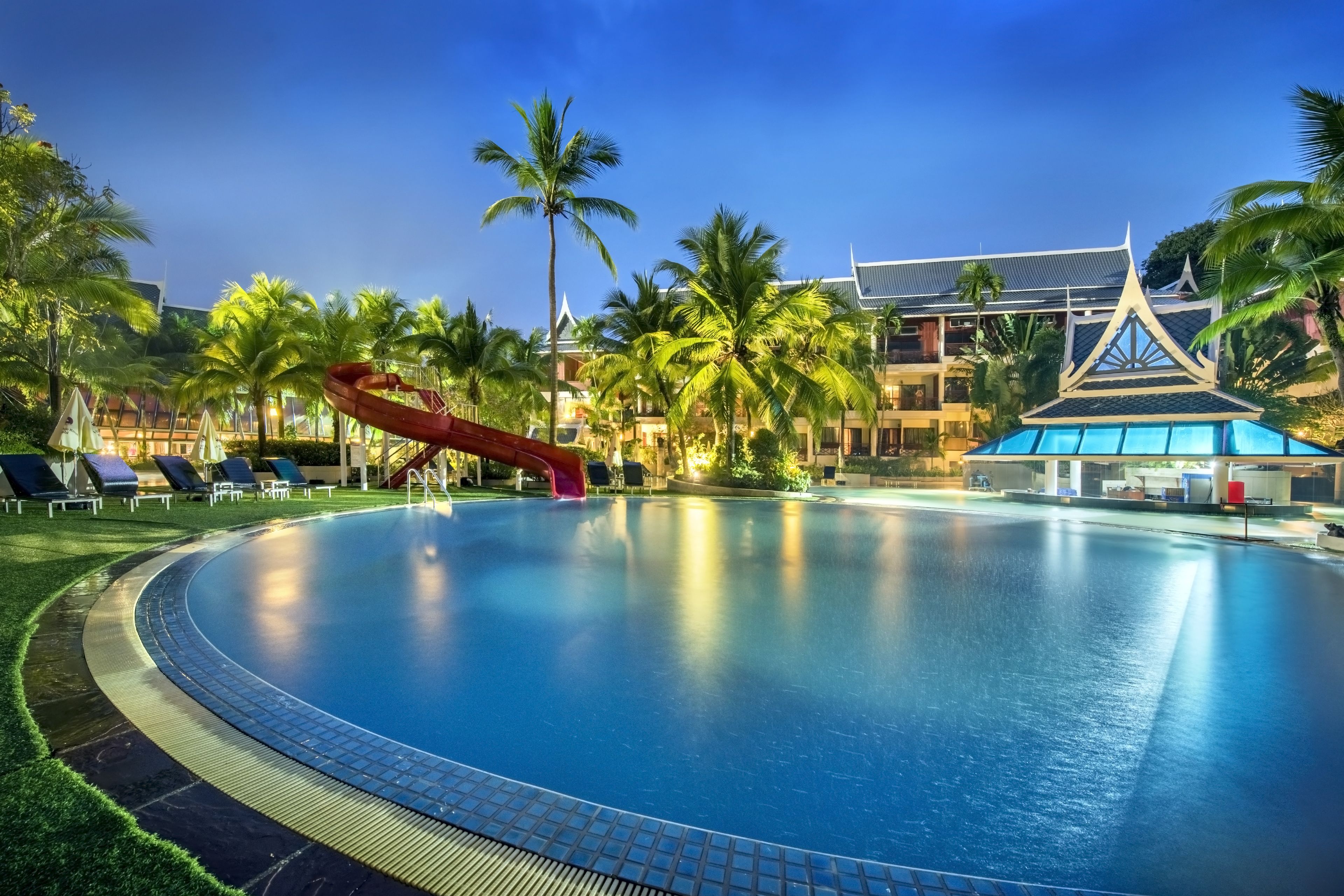A large swimming pool with a water slide, surrounded by palm trees and lounge chairs.
