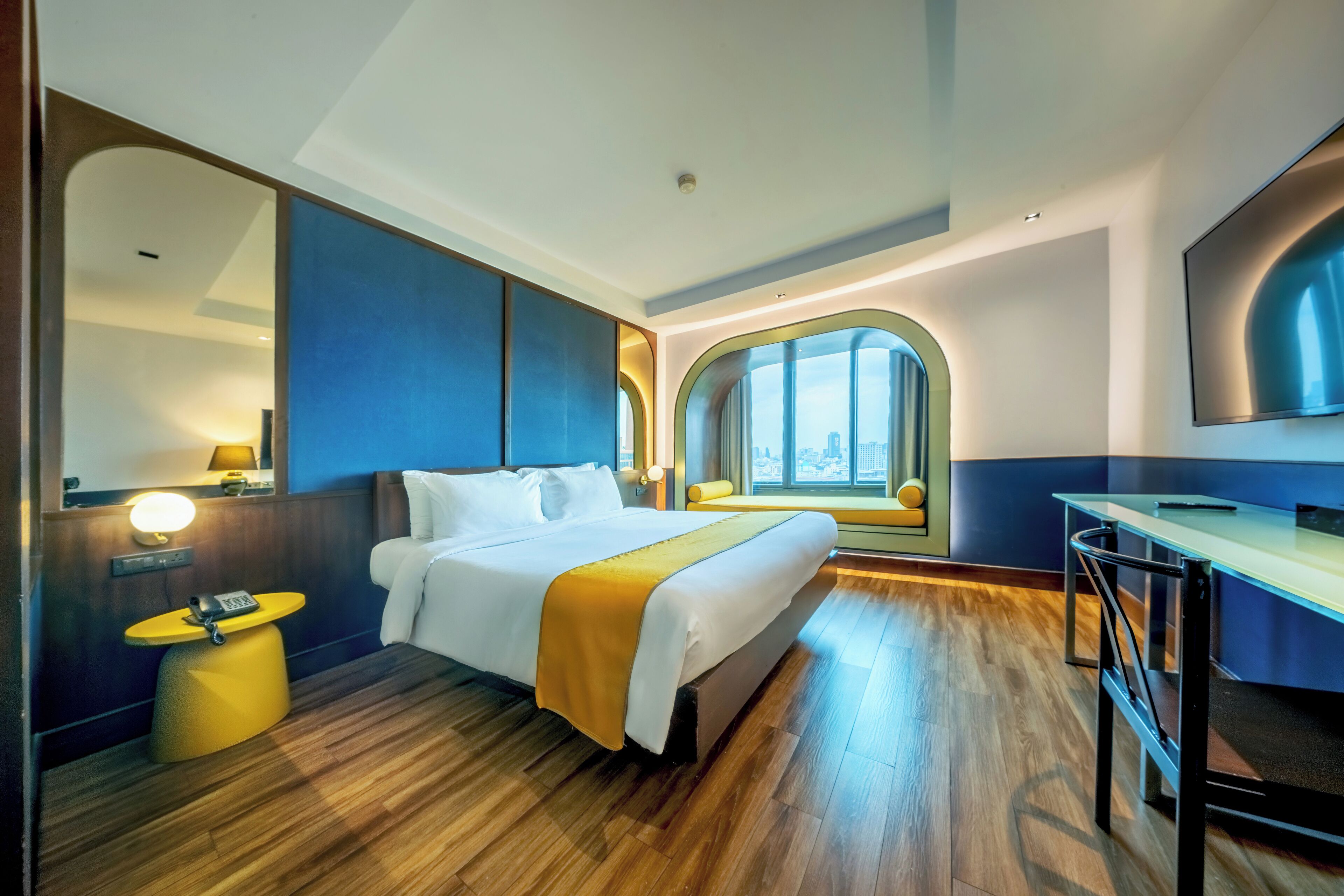 A modern hotel room with a large bed, a desk, a mirror, and a window with a view of the city.