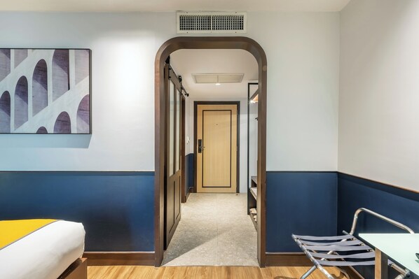 A hotel room with a bed, a desk, a chair, a door, and a hallway leading to another room.