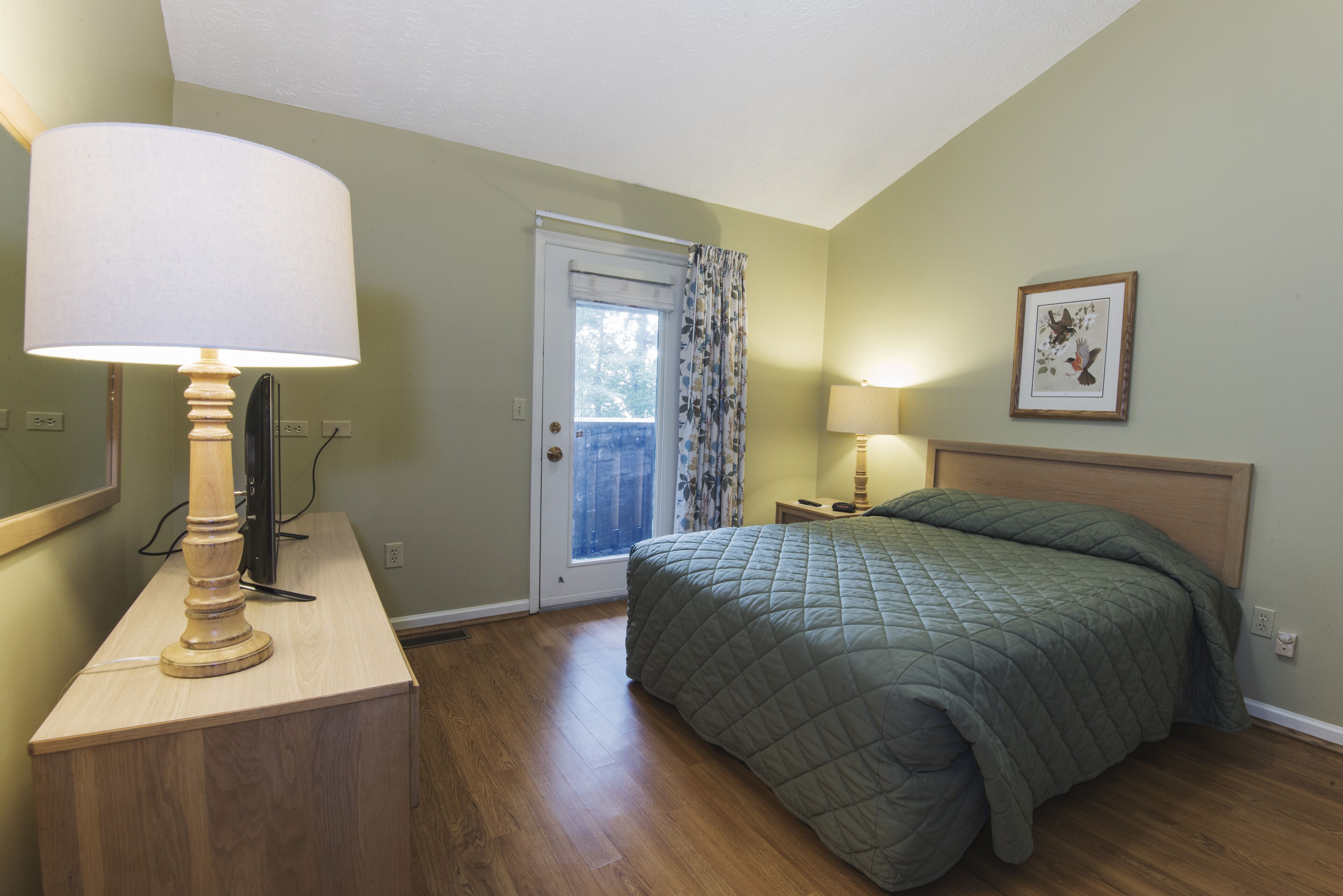 A bedroom with a bed, a desk with a lamp, a door with a view, and a framed picture on the wall.
