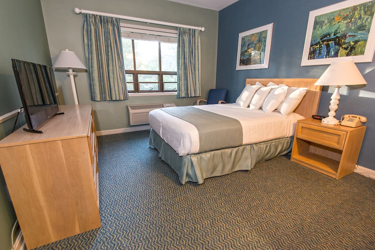 A hotel room with a bed, bedside tables, a desk, a chair, a window with curtains, and artwork on the walls.