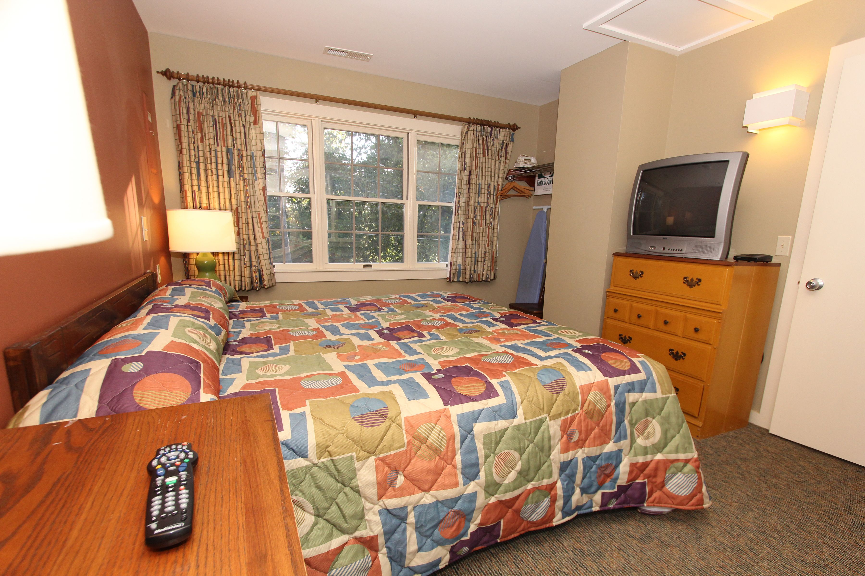 A hotel room with a large bed, a TV on a dresser, a window with curtains, and a lamp on a bedside table.