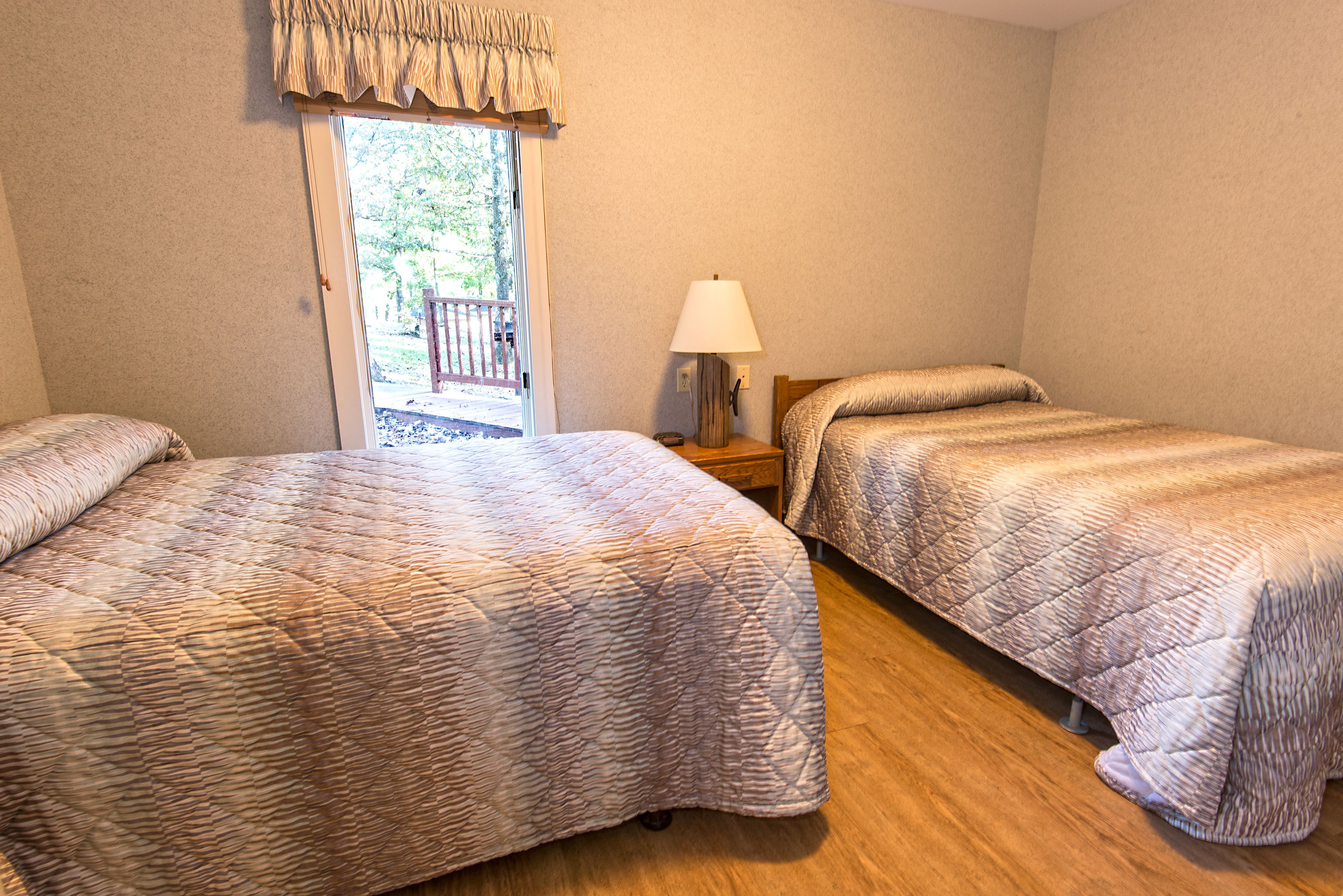 A room with two beds, a wooden floor, a nightstand with a lamp, and a view of the outdoors through a door.