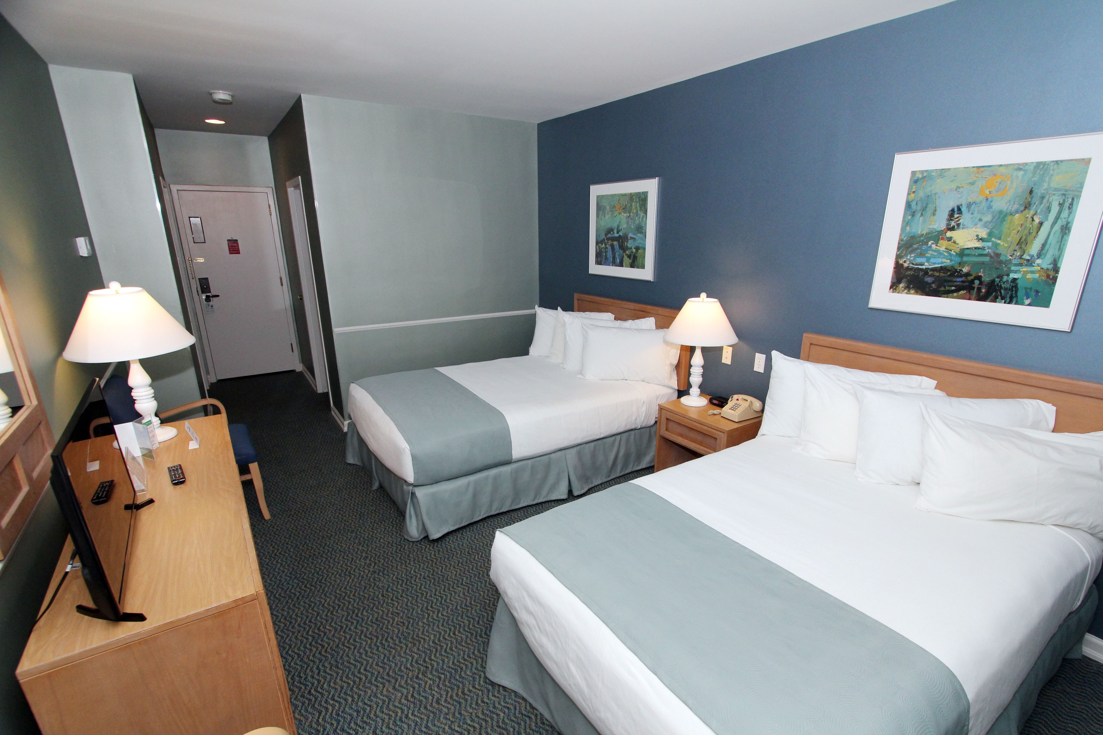 A hotel room with two beds, a desk, and a chair. There are two paintings on the wall, a door, and a carpeted floor.