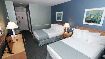 A hotel room with two beds, a desk, and a chair. There are two paintings on the wall, a door, and a carpeted floor.