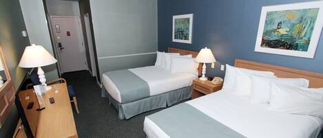Ett hotellrum med två sängar, ett skrivbord och en stol. På väggen hänger två tavlor, och det finns en dörr och ett rum med heltäckningsmatta.