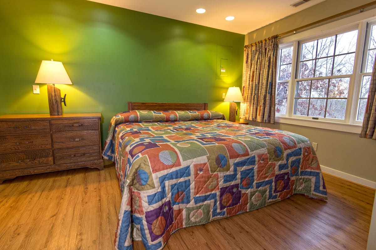 A bedroom with a wooden floor, a green wall, a bed with a colorful quilt, two bedside lamps, and a window with curtains.