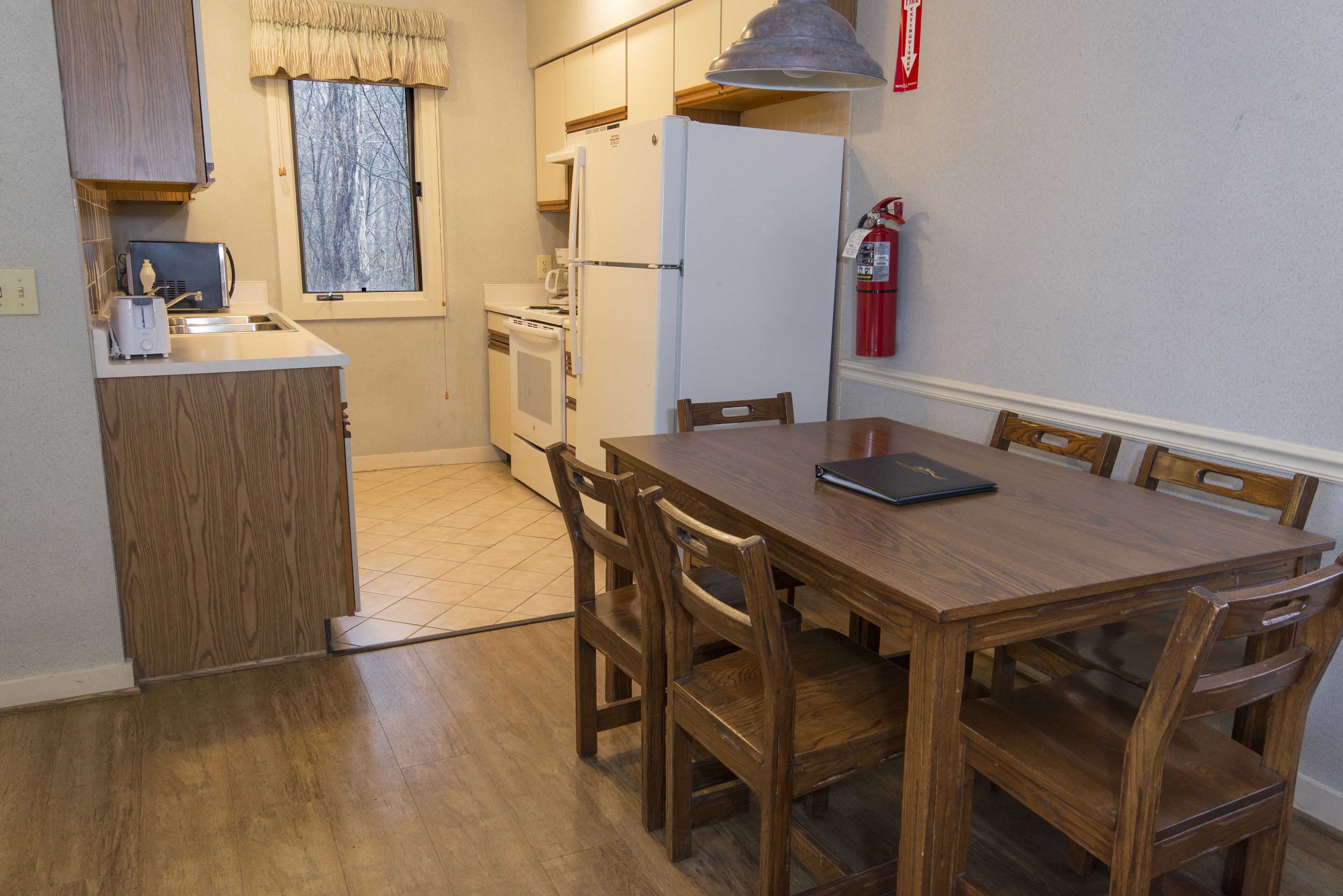 Una cocina compacta con una mesa y sillas, una ventana con cortinas, un refrigerador blanco y un extintor en la pared.