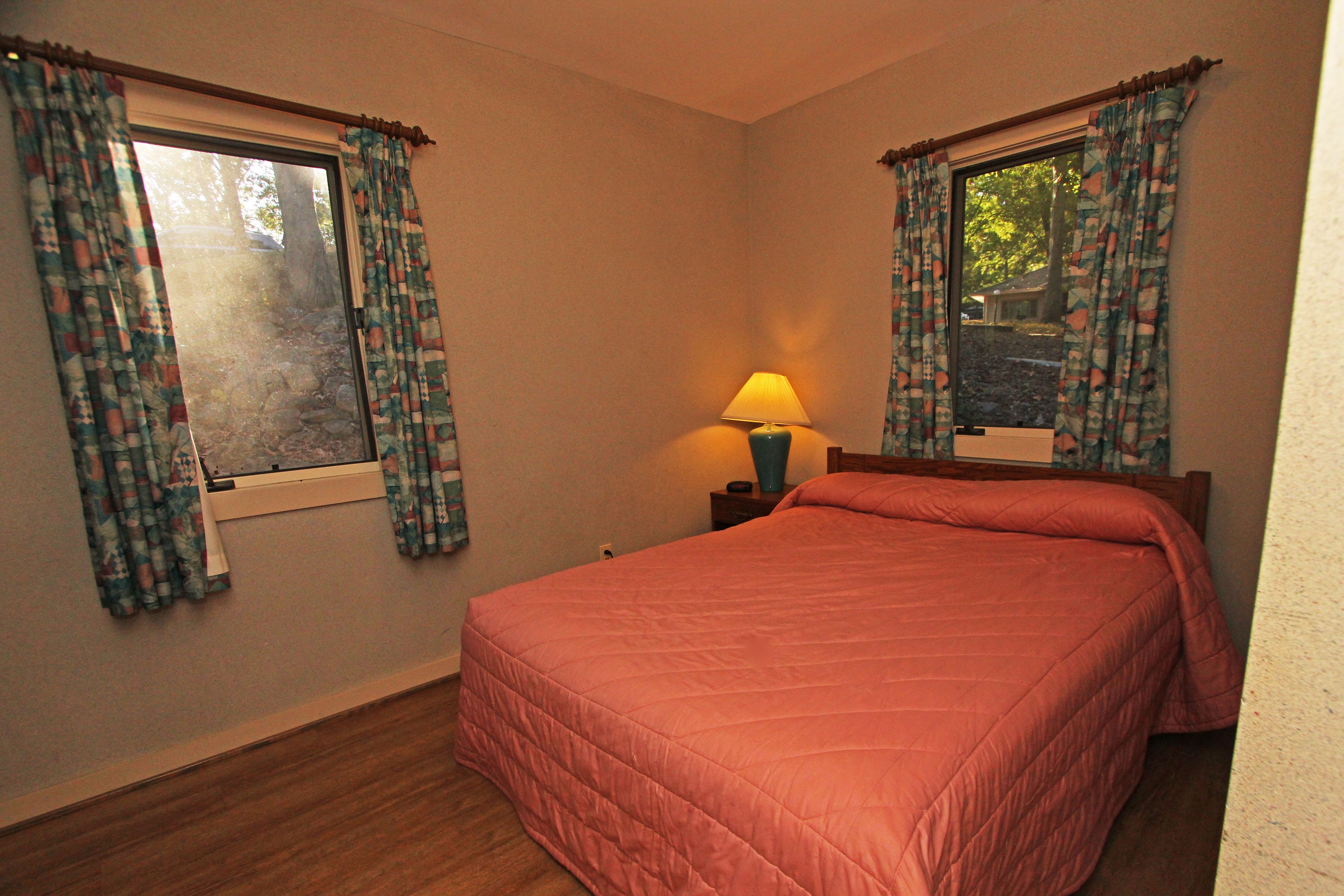 Une chambre à coucher avec un lit, deux fenêtres avec des rideaux, une table de chevet avec une lampe, et une vue sur l’extérieur.