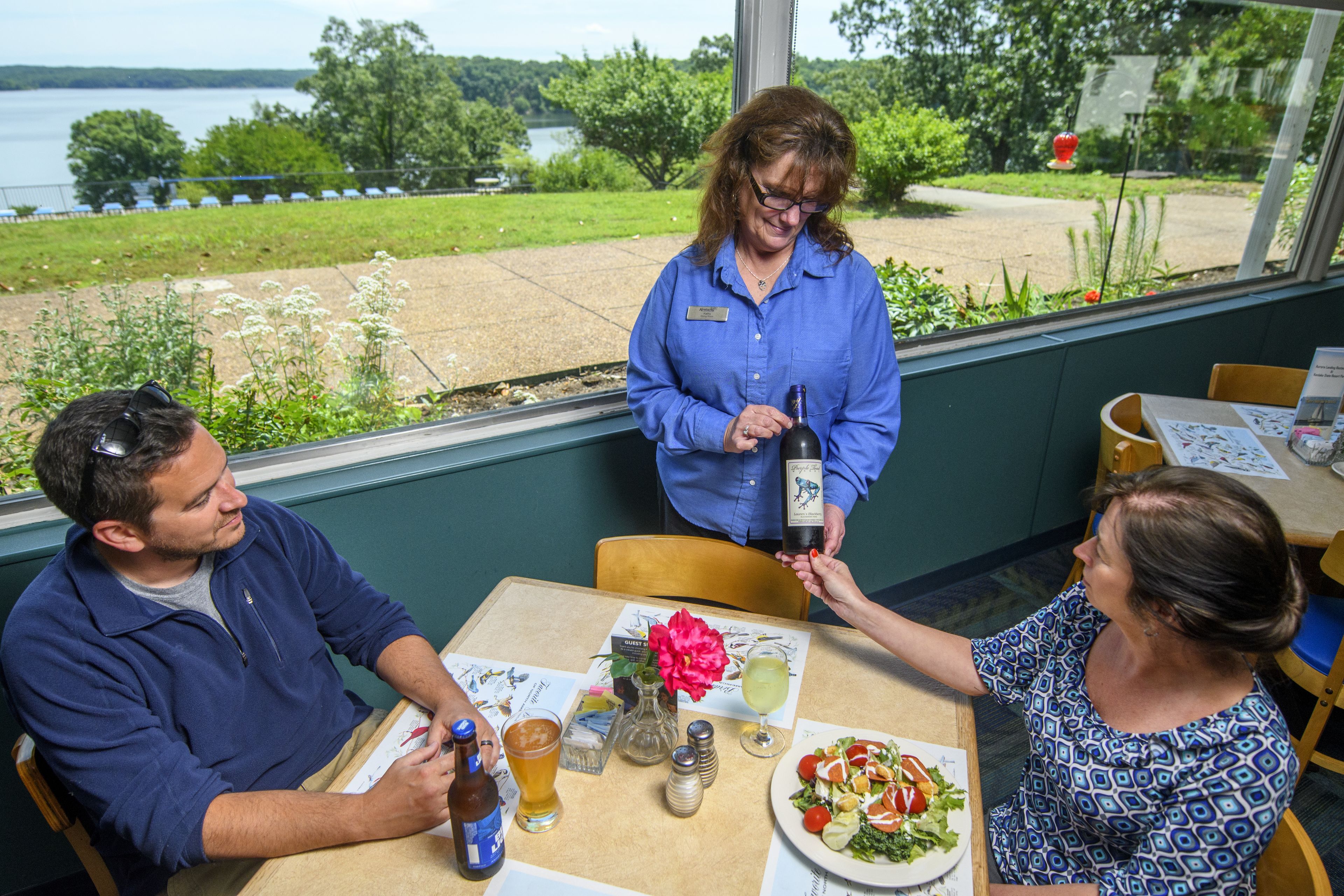 A restaurant with a view of a lake, a table with a meal, and a person holding a wine bottle.