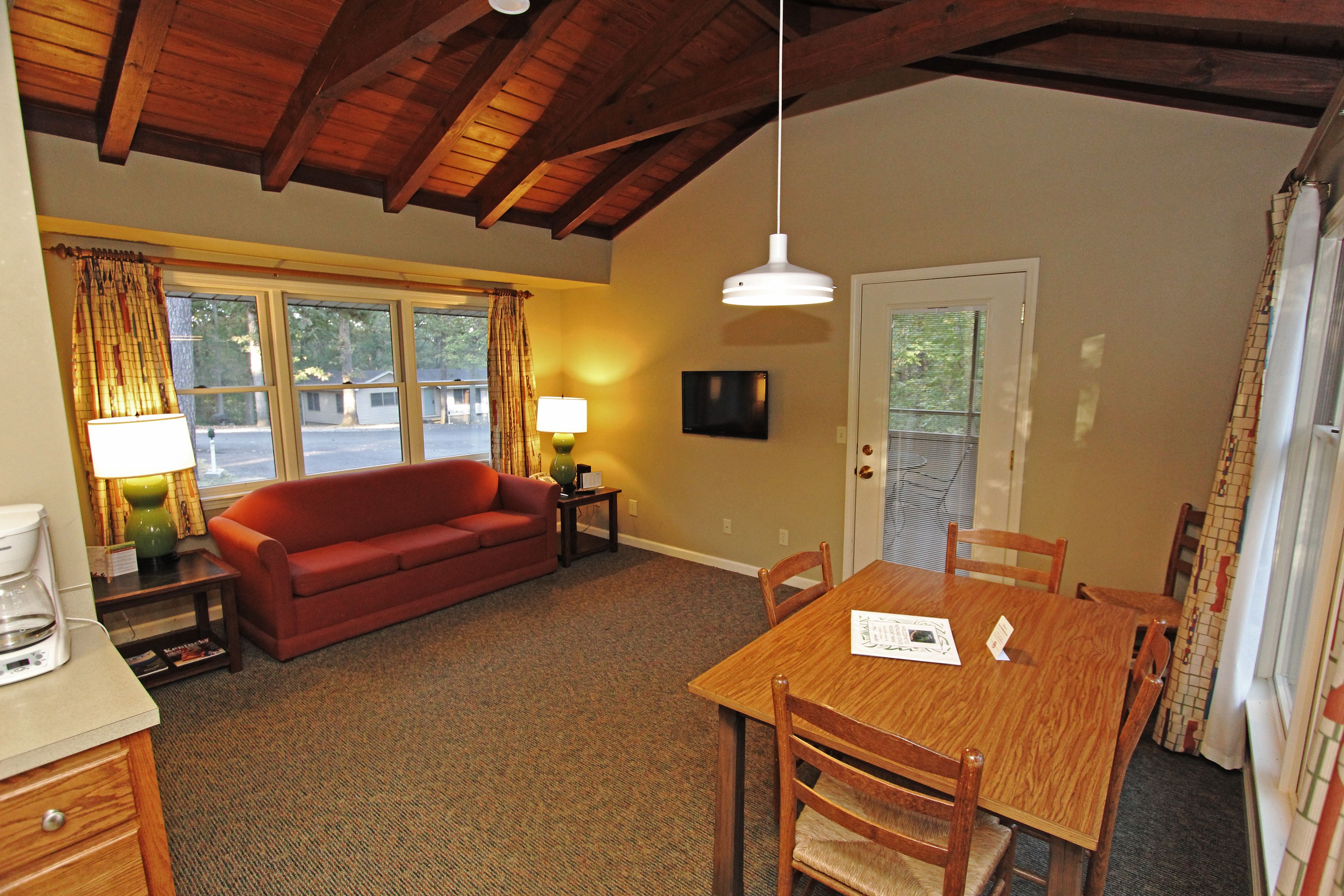 A cozy living room with a red sofa, wooden dining table, and chairs, a coffee maker, and a lamp on a side table.