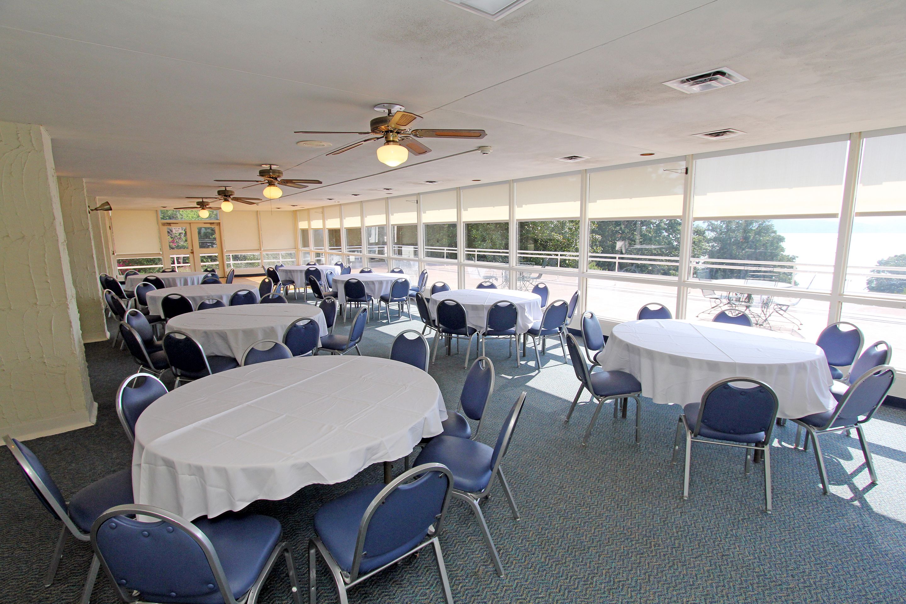 Une salle de conférence dotée de tables et de chaises rondes, de grandes fenêtres et de ventilateurs de plafond.
