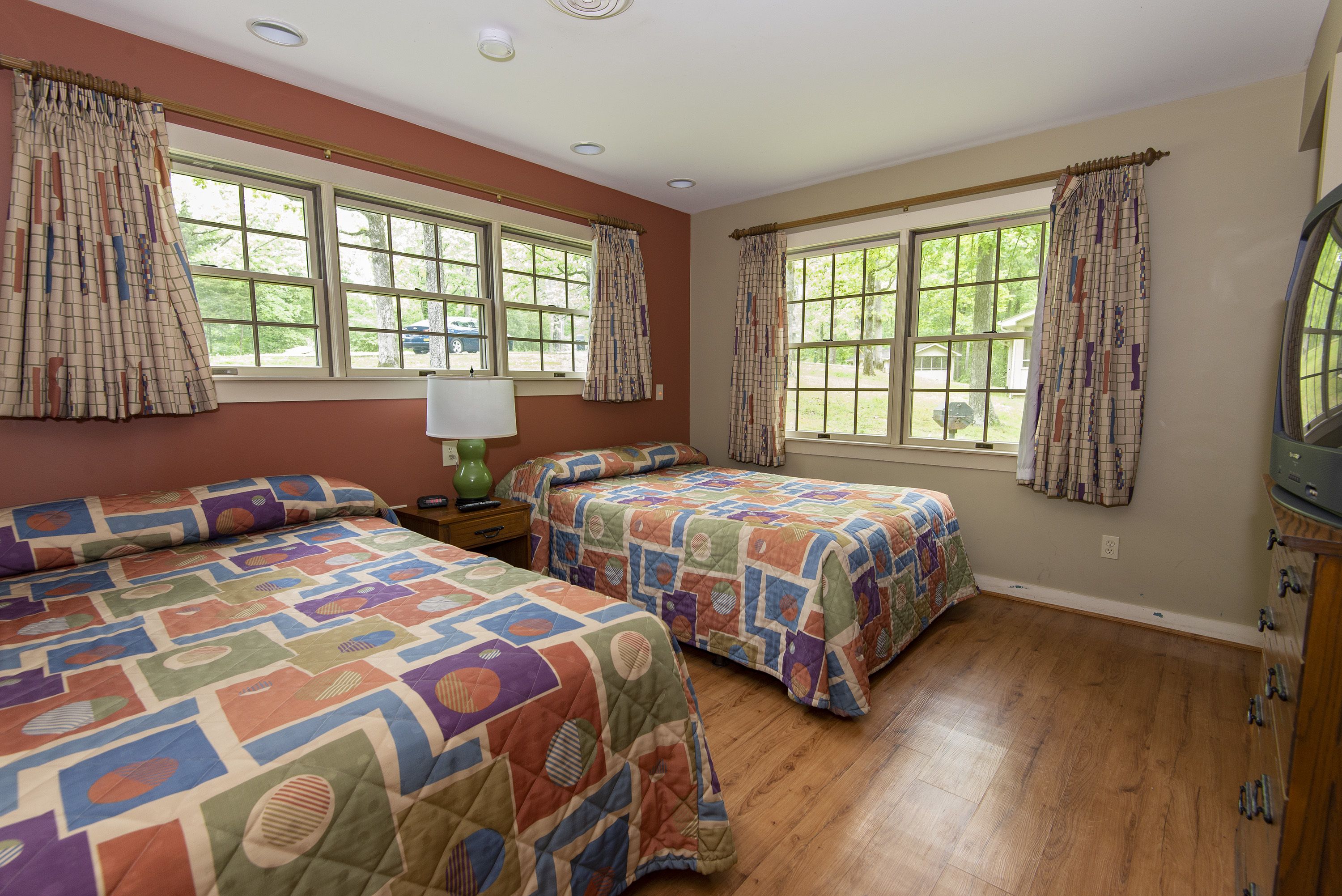 A bedroom with two beds, large windows, a nightstand with a lamp, and a dresser.