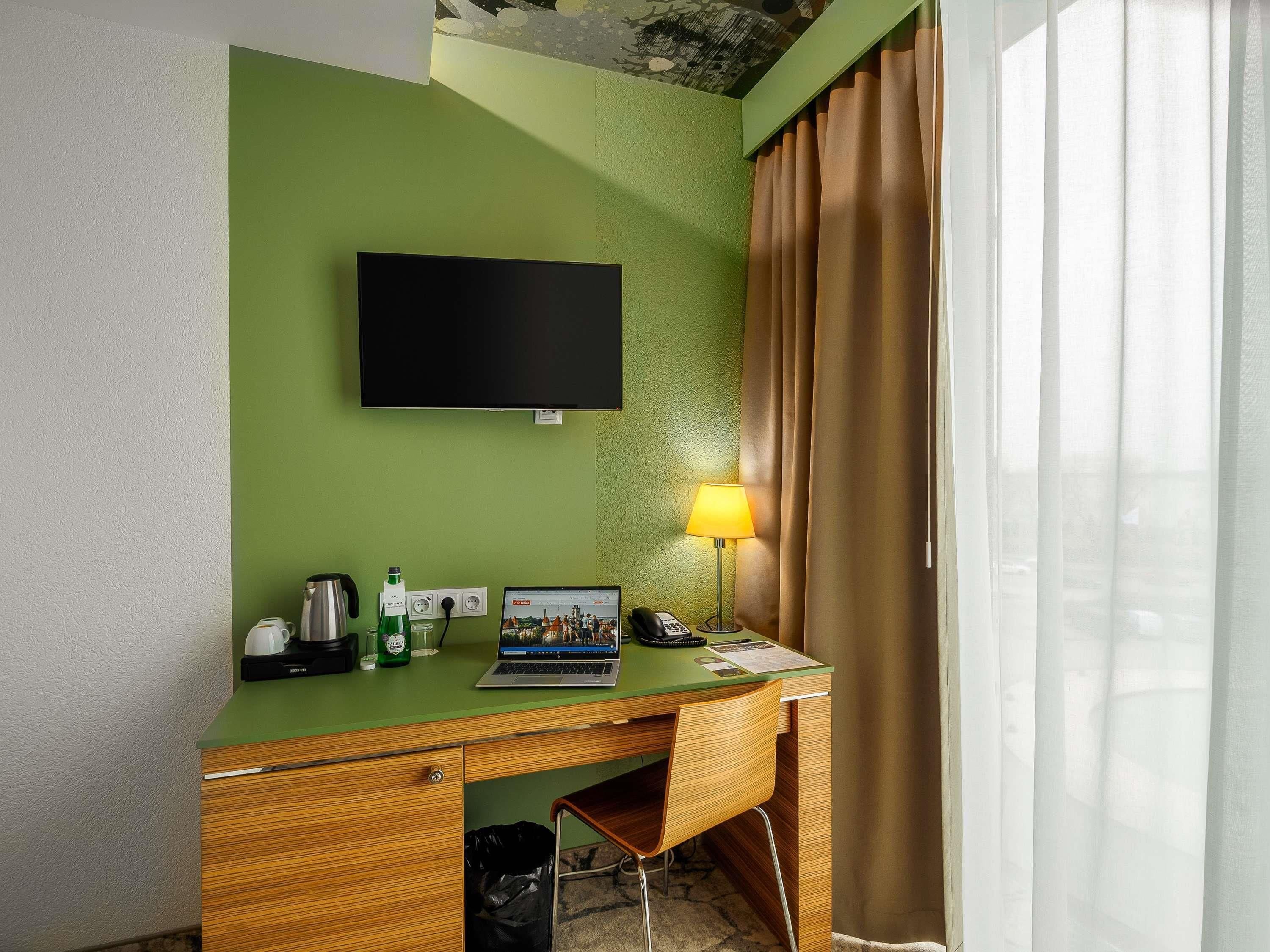 Habitación de hotel con pared verde, televisor de pantalla plana, escritorio con ordenador portátil, lámpara y cafetera.