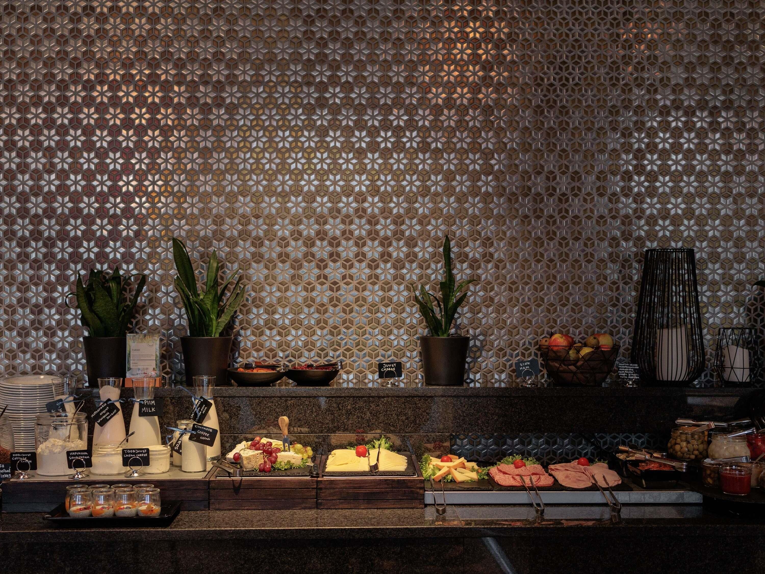 Mesa de buffet con diversos alimentos y bebidas, incluyendo queso, frutas y salsas, frente a una pared con diseño.
