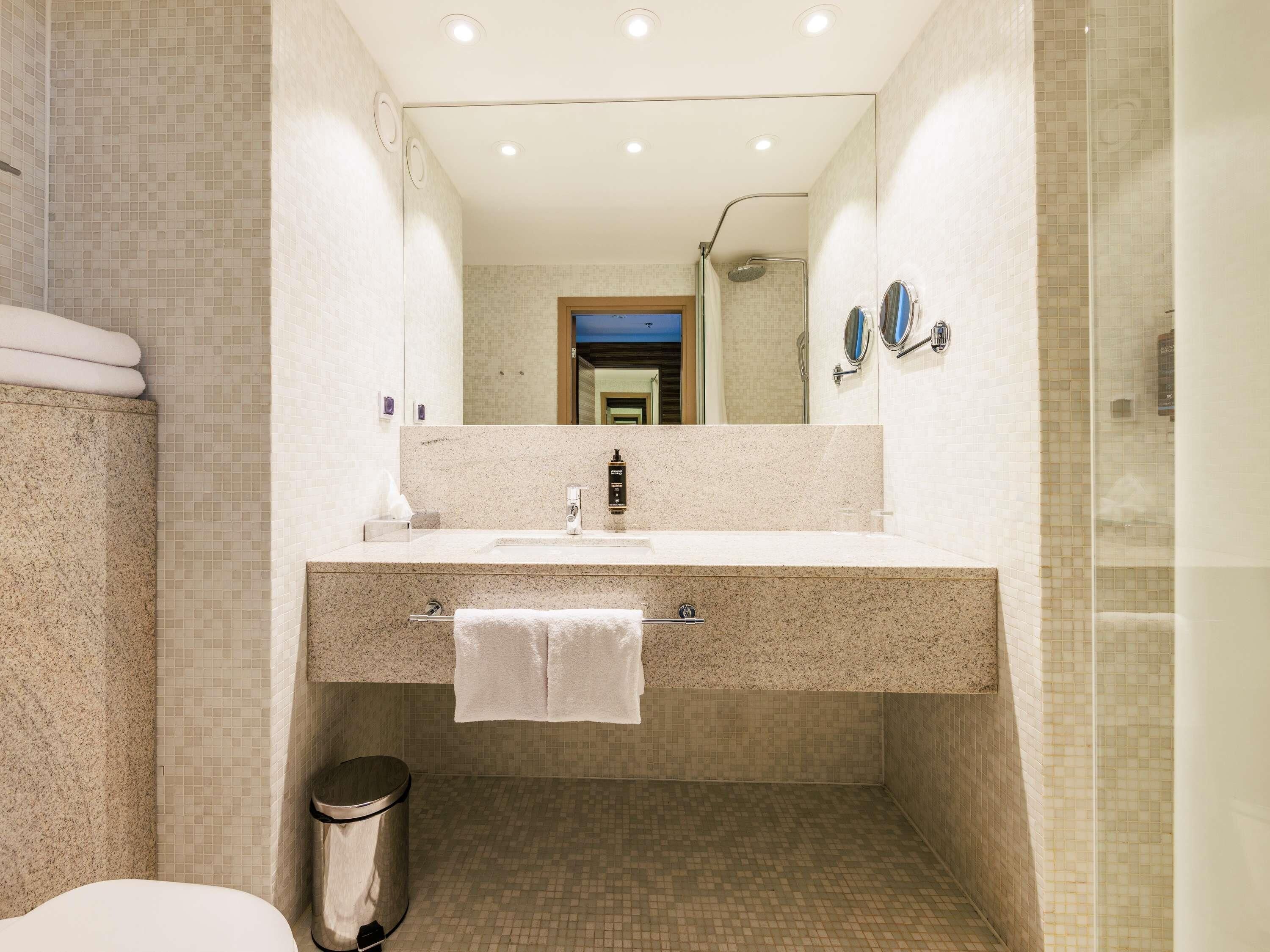 Un baño con lavamanos, espejo, toallero y cubo de basura.