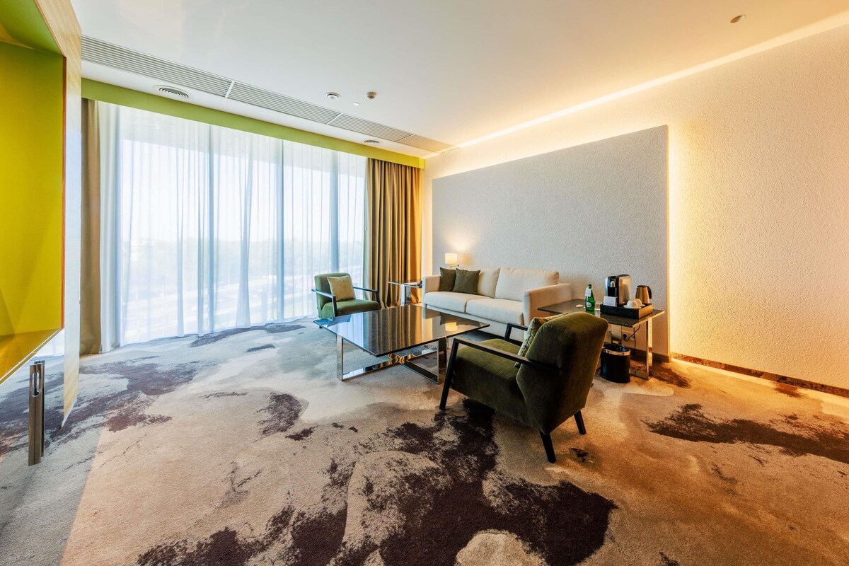 A hotel room with a large window, a sofa, a chair, a coffee table, and a desk with a lamp.
