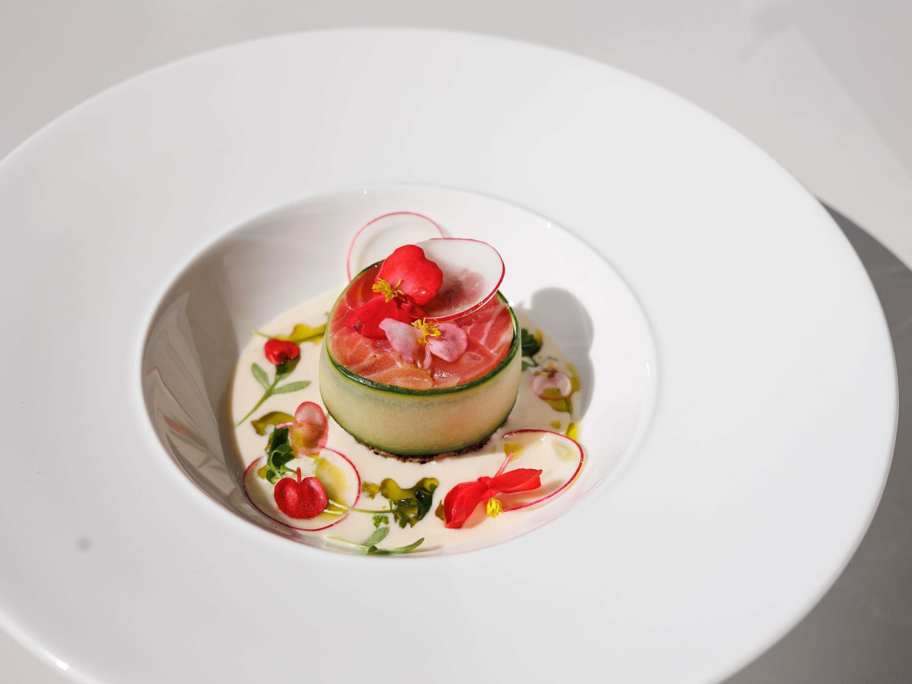 A white plate with a layered dish, garnished with red flowers and thin slices of fruit.