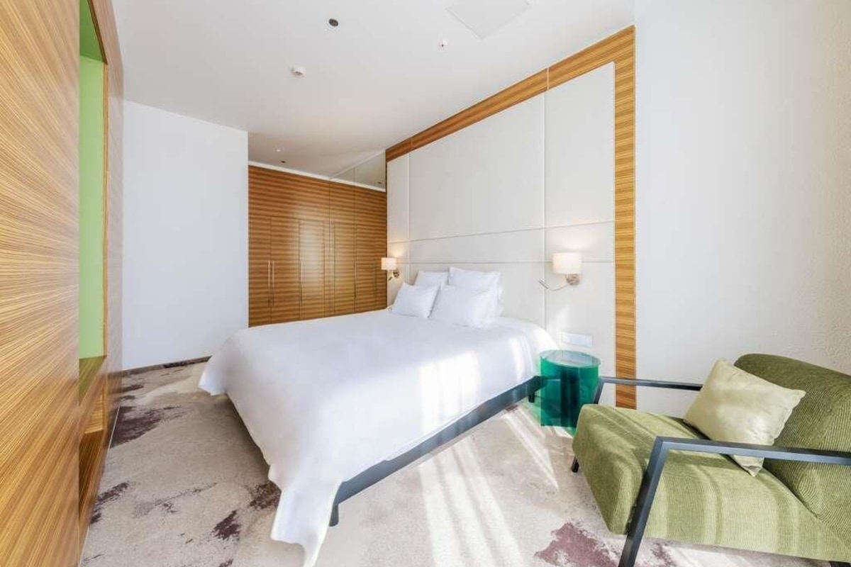A modern hotel room with a large bed, a green armchair, a glass side table, and a wooden accent wall.