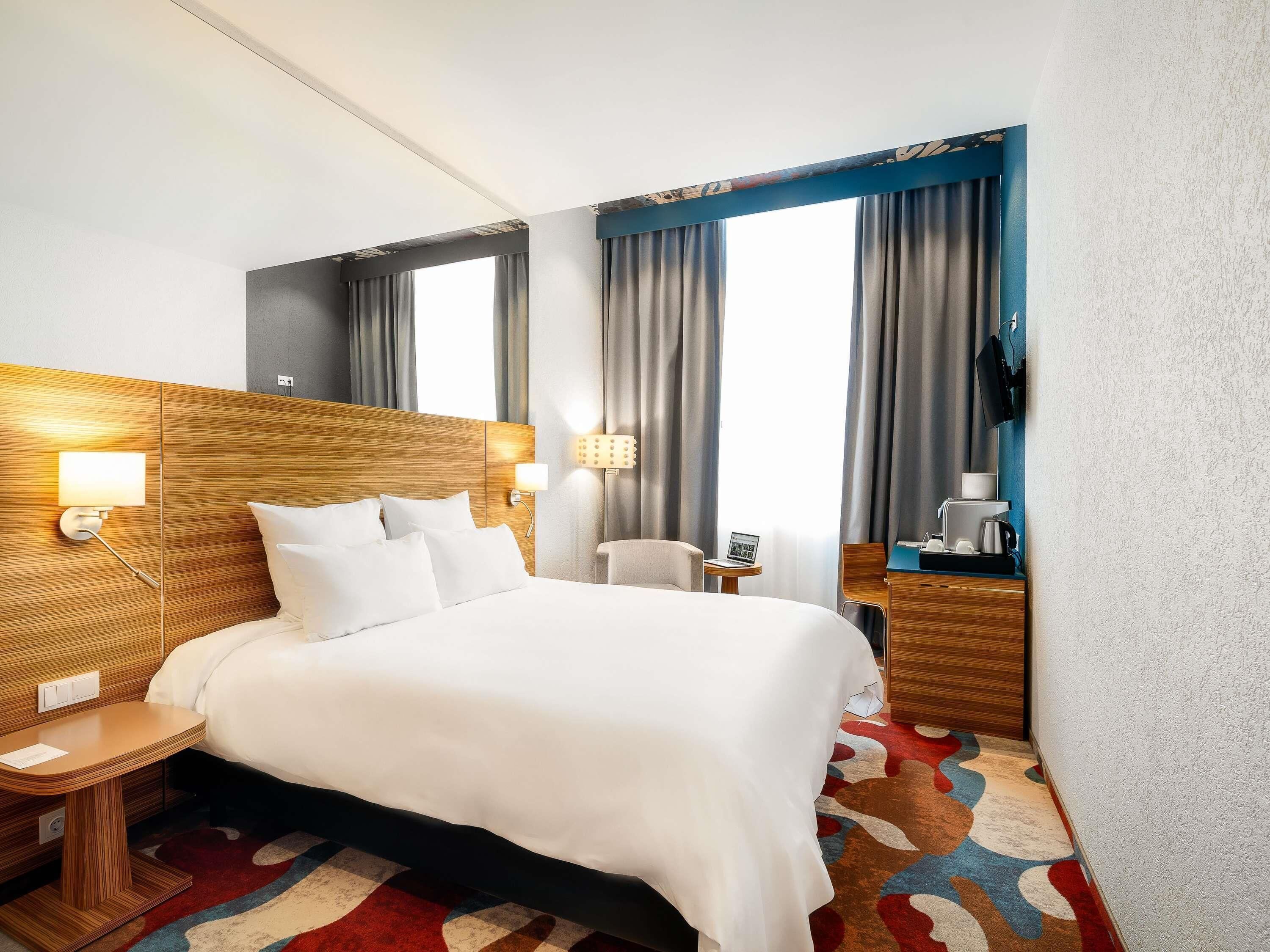 Une chambre d’hôtel avec un grand lit, des tables de chevet, un bureau, une chaise, une télévision et une fenêtre avec des rideaux.